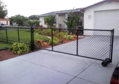 Black chain-link fence with rolling gate in front of a house with a driveway, and a lush garden.