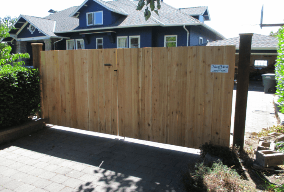Wooden double gate on a brick driveway, leading to a blue house.