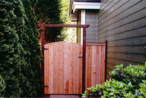 Wooden gate and fence next to green siding and bushes, with a green bush to the left.