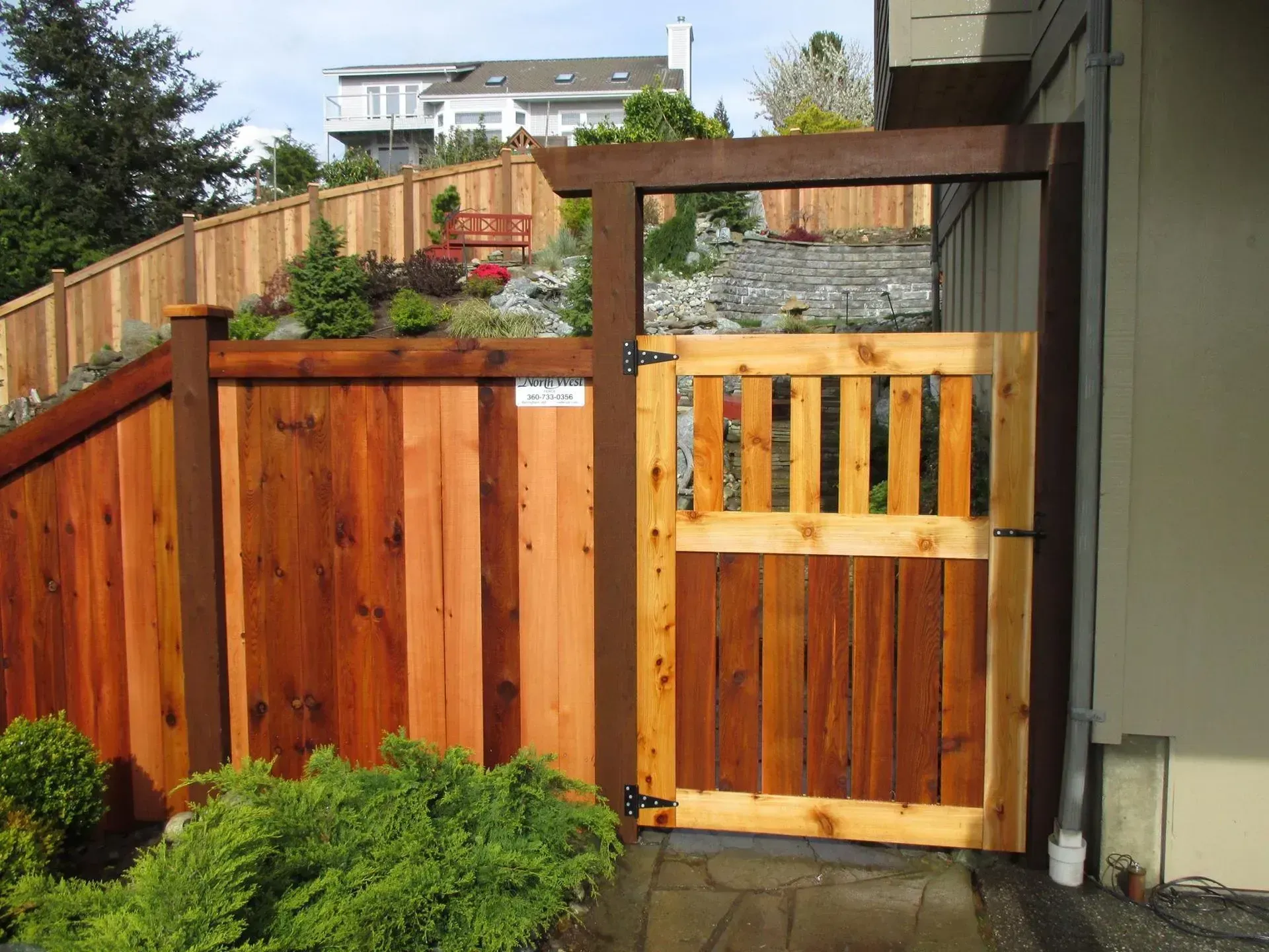 Wooden fence with gate leading to a backyard, brown and reddish wood, shrubs in front.