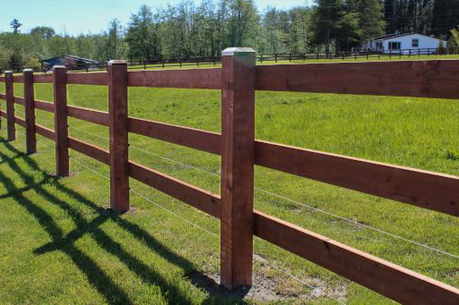 Wooden fence with three horizontal rails, enclosing a green field, with trees and a house in the background.
