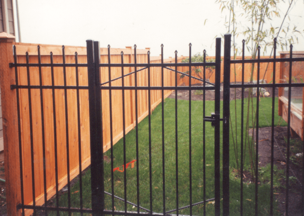 Black wrought iron gate with a wooden fence and green grass in the background.