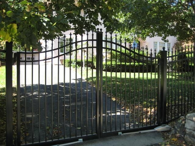 Black metal driveway gate with curved top, open, leading to a house with green lawn and trees.