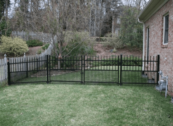 Black metal fence surrounding a grassy yard near a brick house and trees.