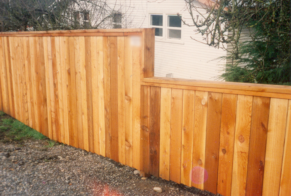 Wooden privacy fence in front of a white house, constructed of vertical wooden planks.
