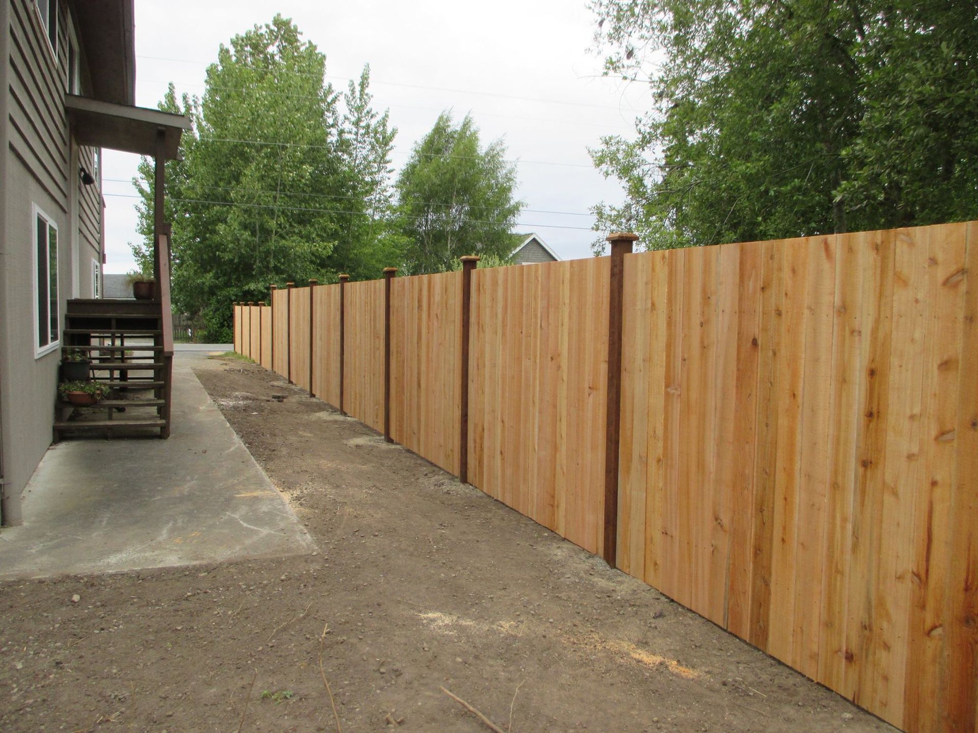 Wooden fence running along a gray building and a gravel path, with trees in the background.