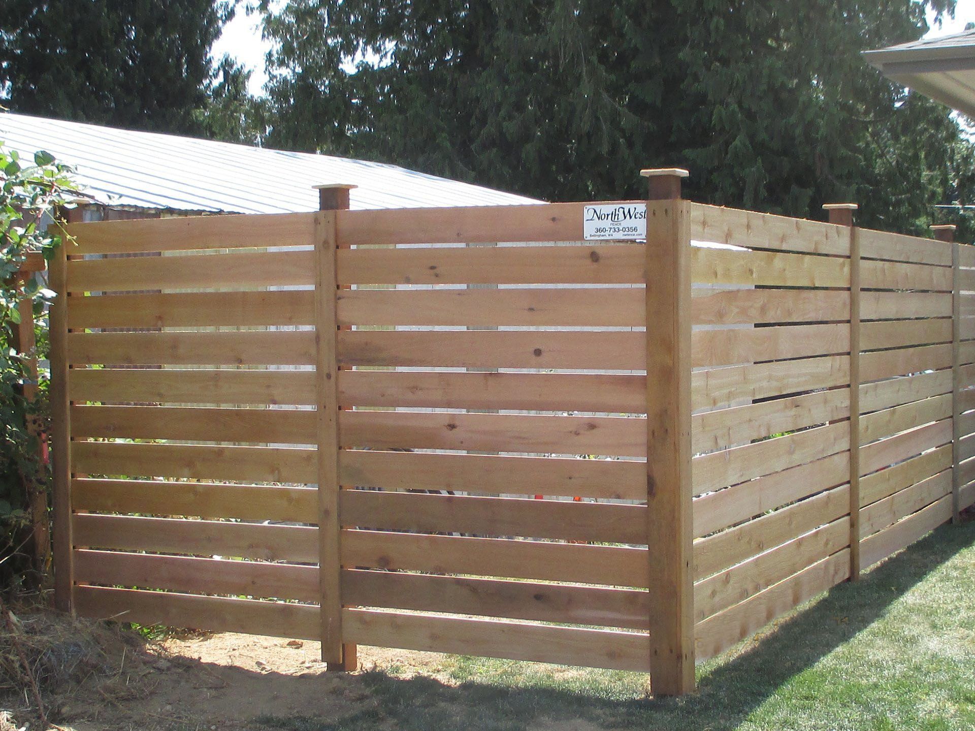 Wooden horizontal slat fence in a backyard with a white building and green grass.