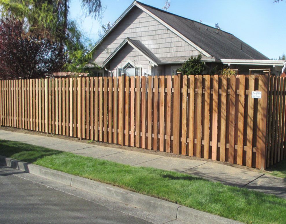 Brown wooden fence surrounding a house with a gray roof, next to a sidewalk and lawn.