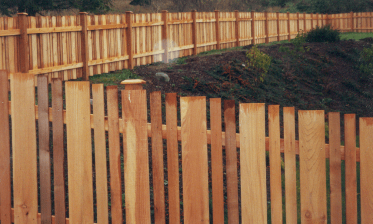 Wooden fence with varying height pickets bordering a grassy area.