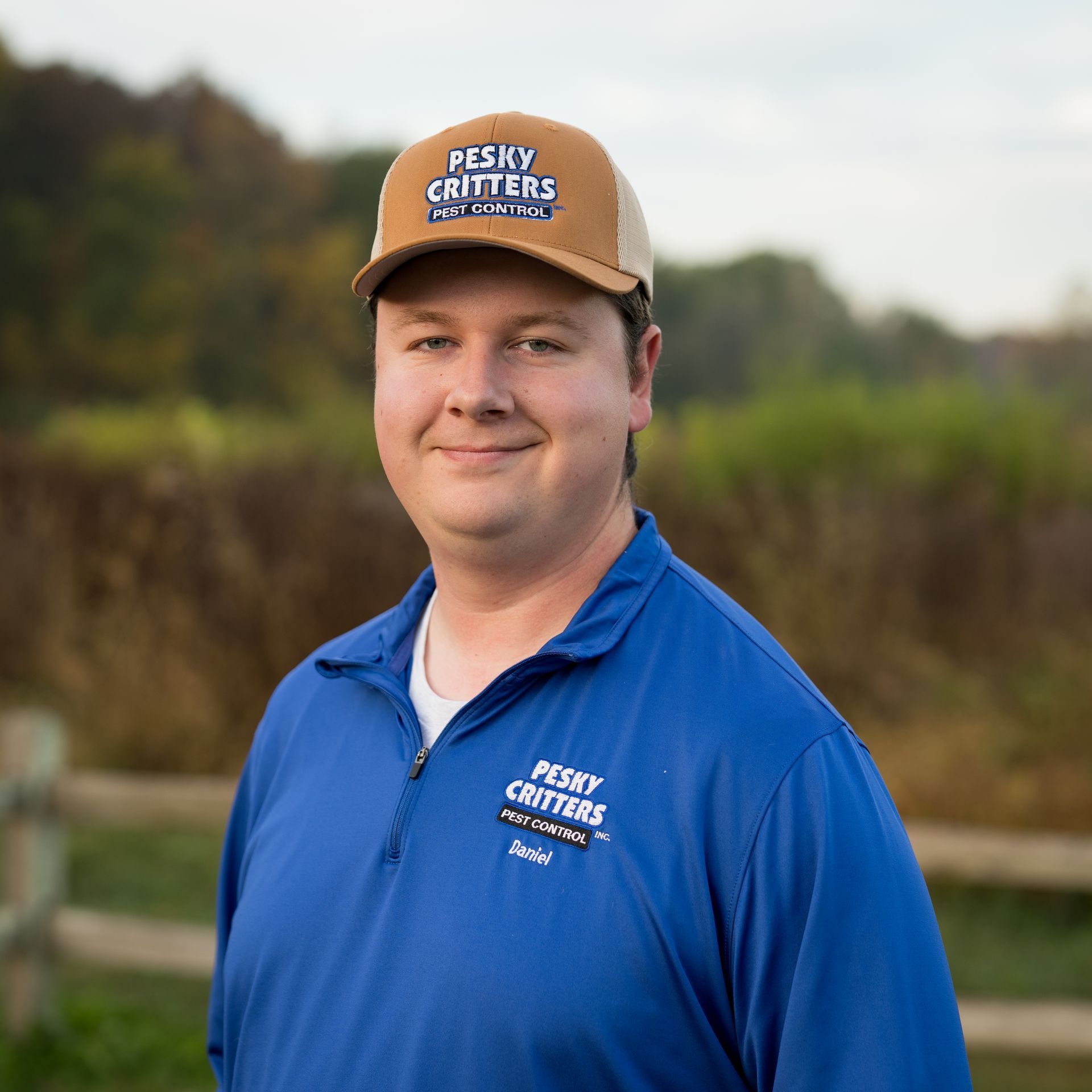 A person wearing a blue branded pullover and a tan trucker hat stands outdoors in front of a blurred field.