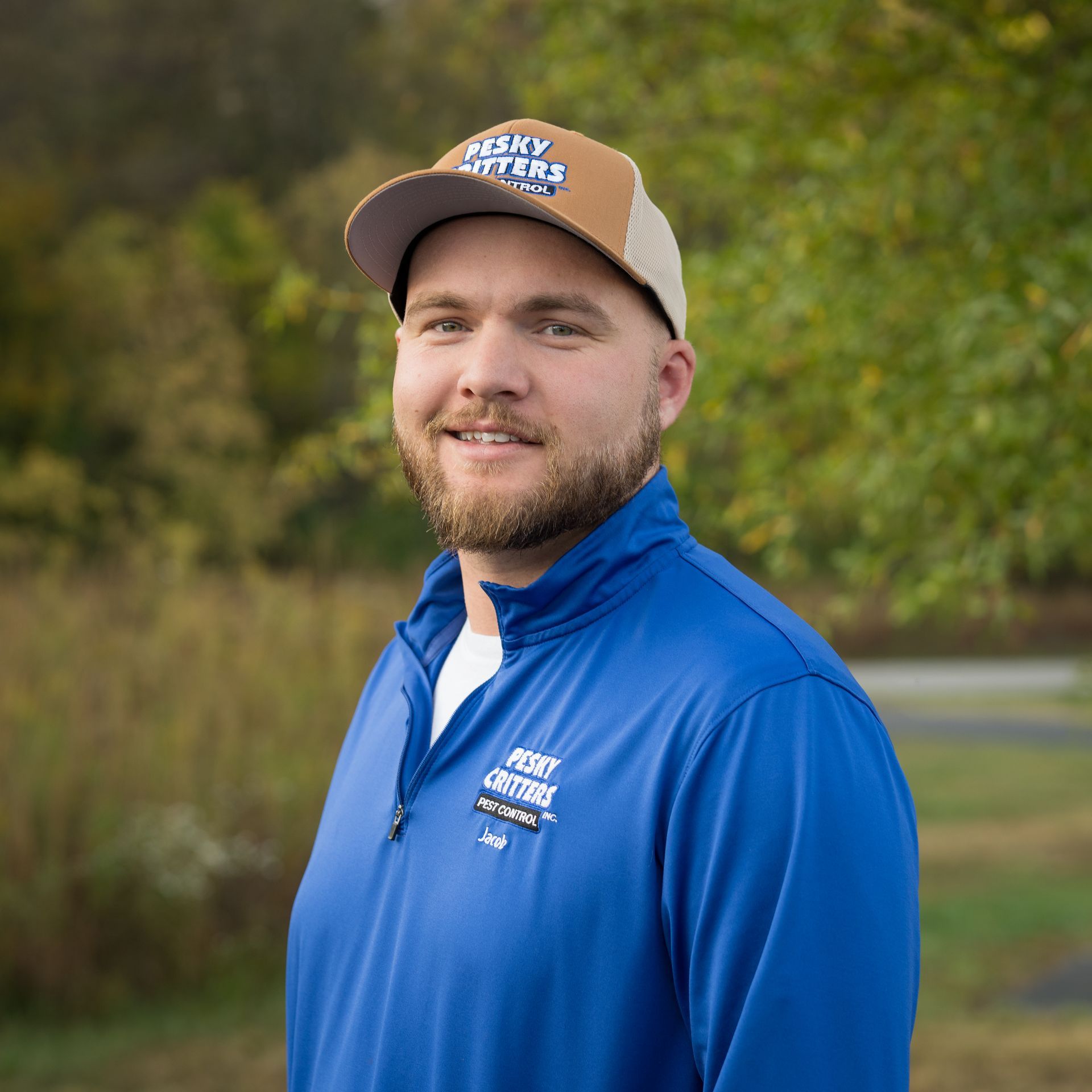 A person wearing a blue zip-up shirt and a tan and brown baseball cap stands outdoors against a blurred nature background.