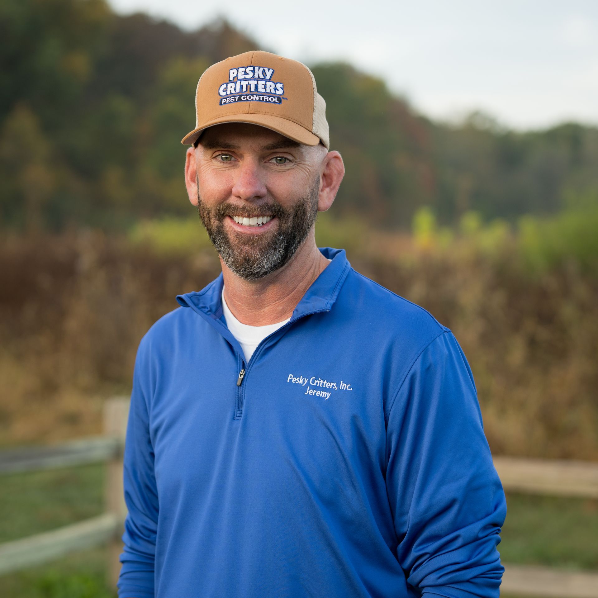 A smiling person wears a tan cap and blue long-sleeve quarter-zip shirt, standing outdoors in front of a field and fence.