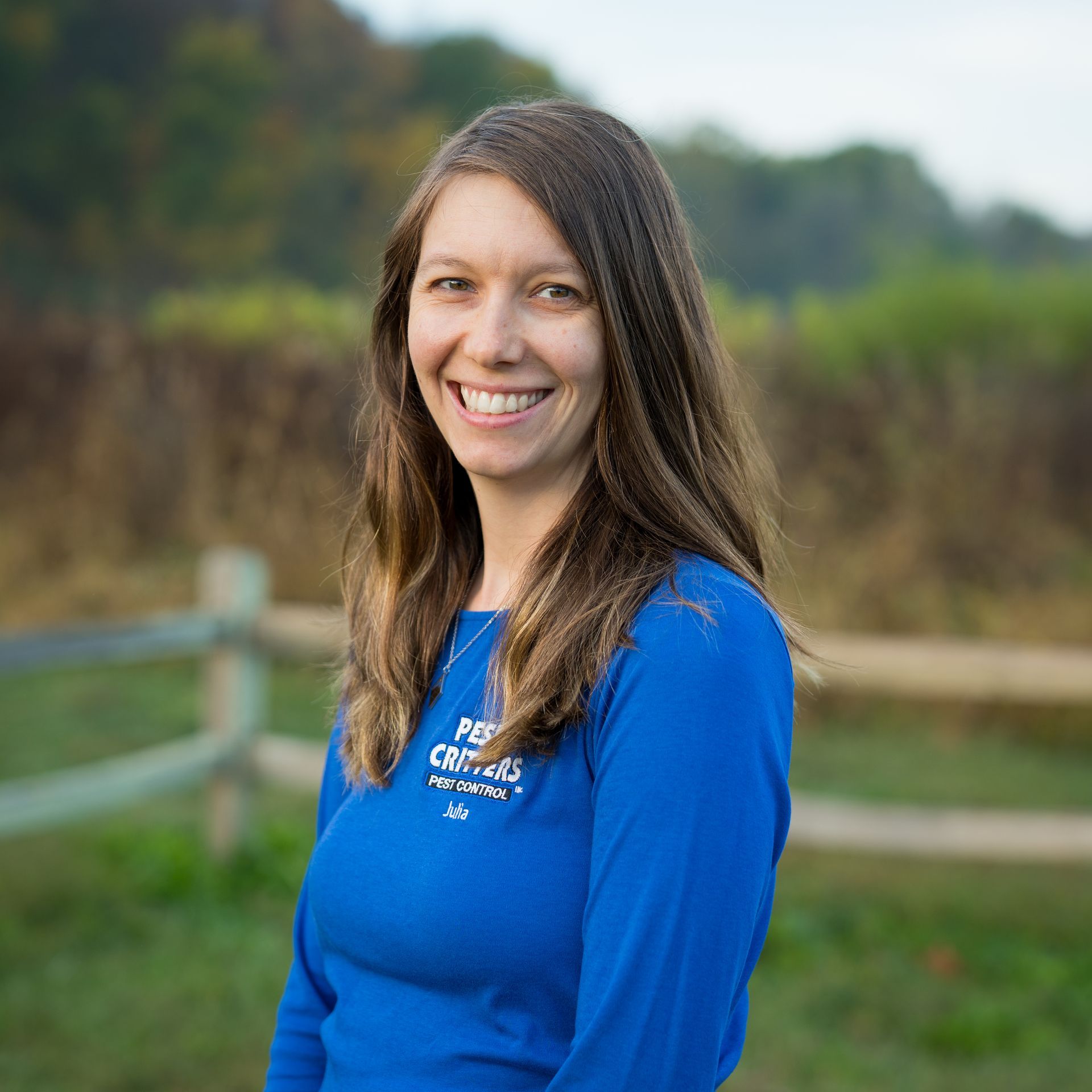 A smiling person wearing a blue long-sleeved shirt with a logo, standing outdoors in front of a wooden fence and field.