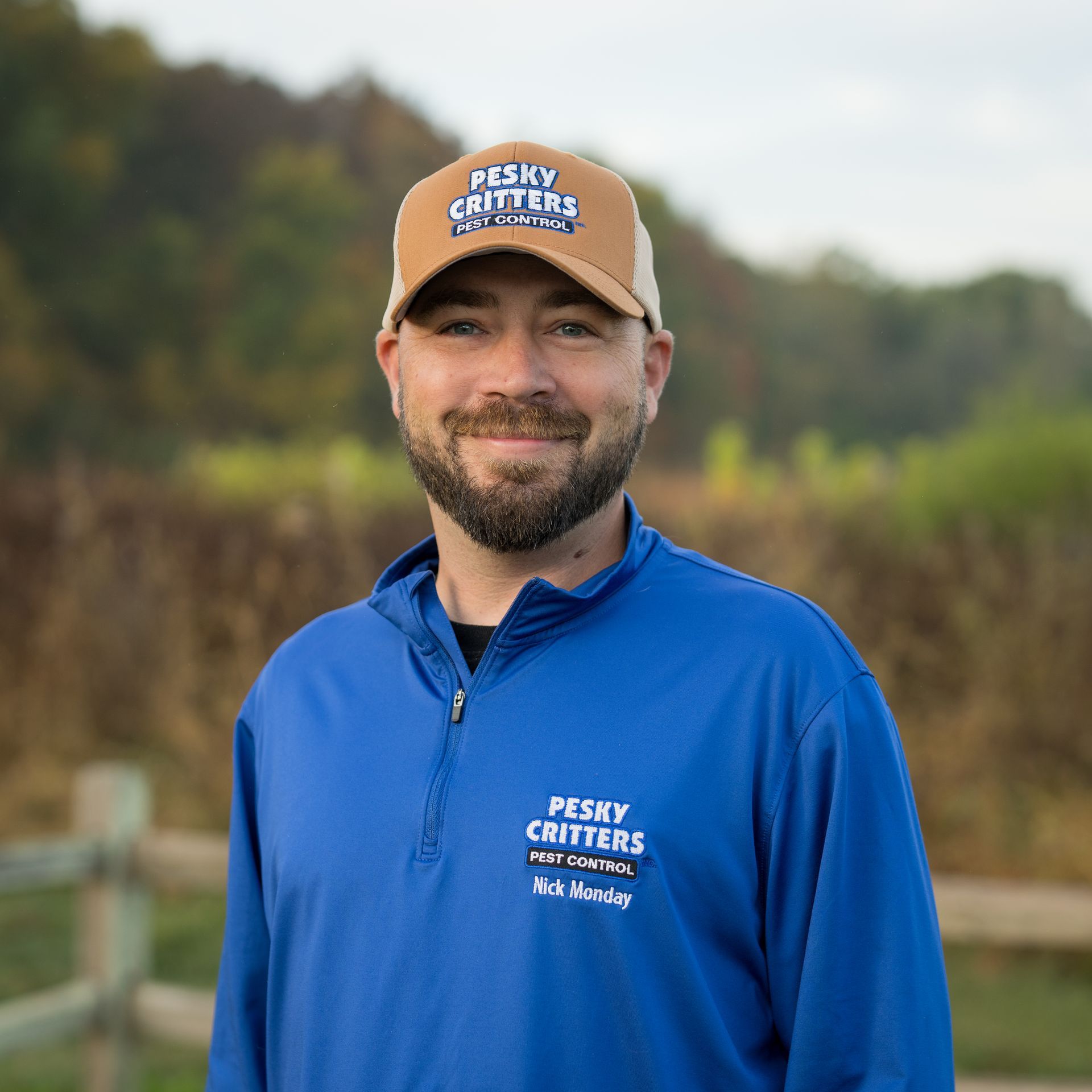 A man with a beard, wearing a brown baseball cap and blue quarter-zip, smiles outdoors in front of a field and fence.