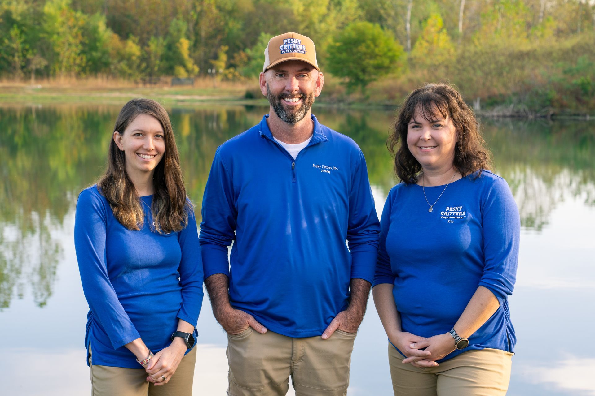 Three people in blue long-sleeve shirts and tan pants stand smiling side-by-side in front of a calm lake.