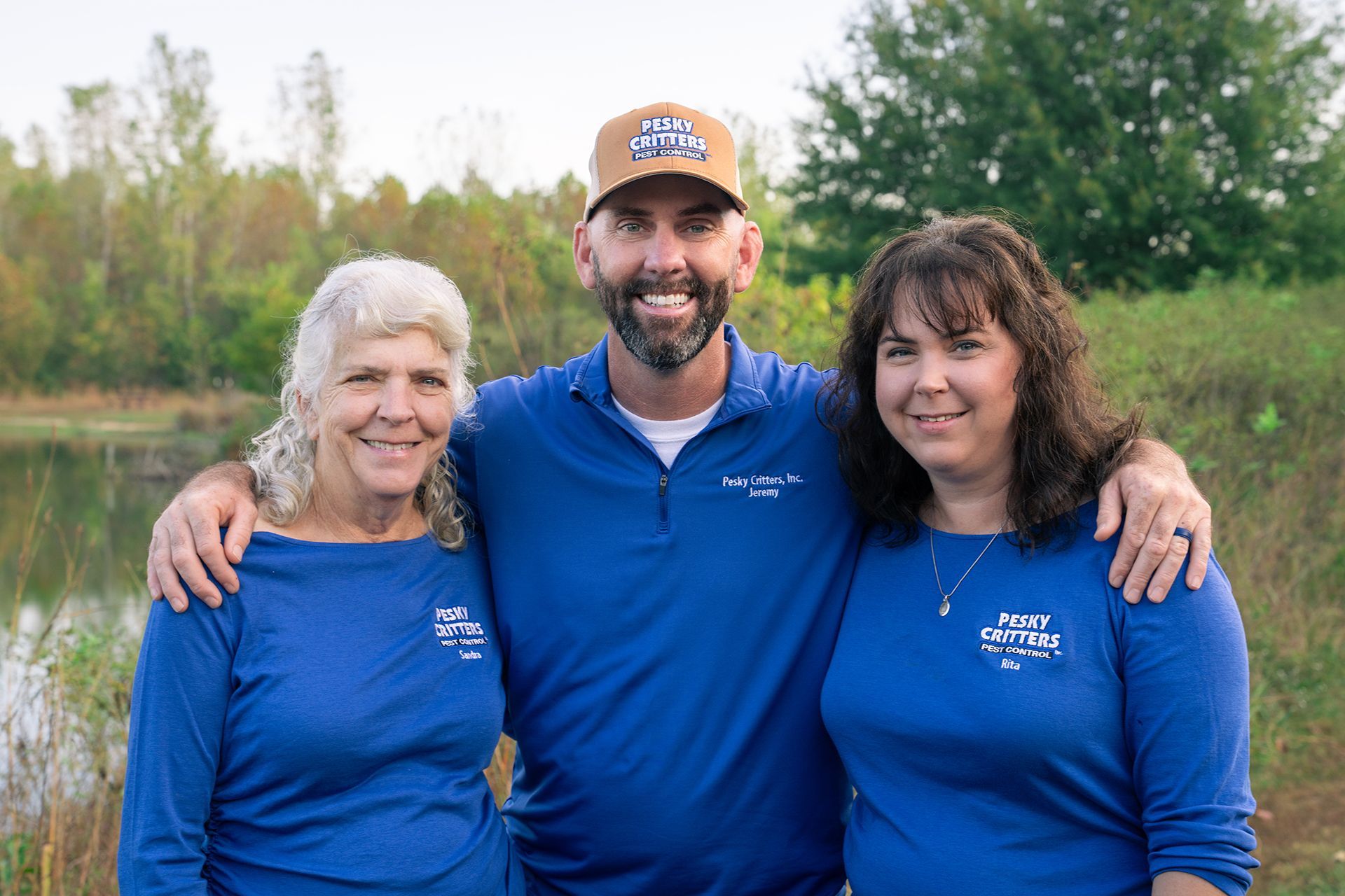 Three people in matching blue shirts pose together outdoors with a pond and trees in the background.