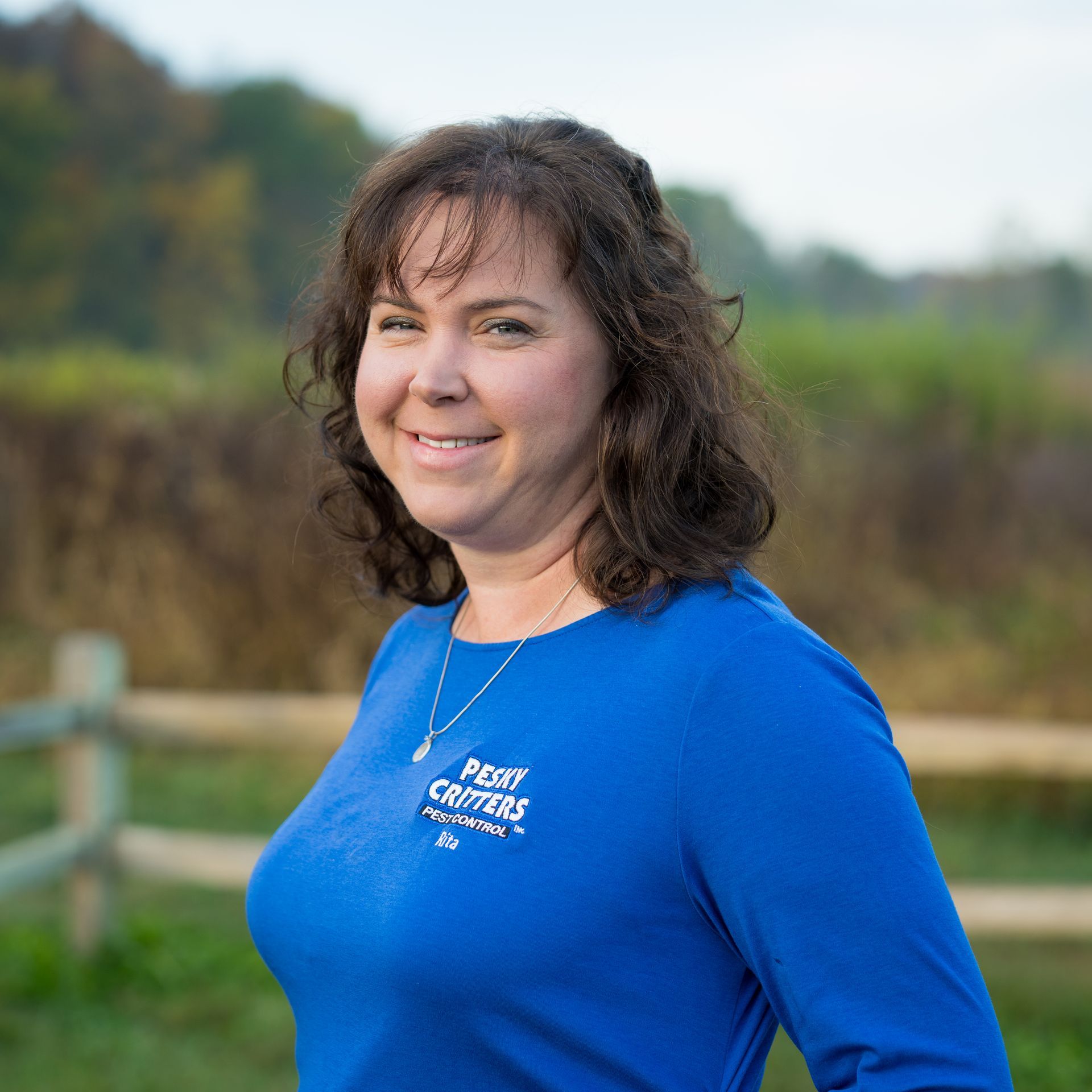 A smiling person wears a blue long-sleeved shirt with a logo, standing outdoors in front of a wooden fence and greenery.