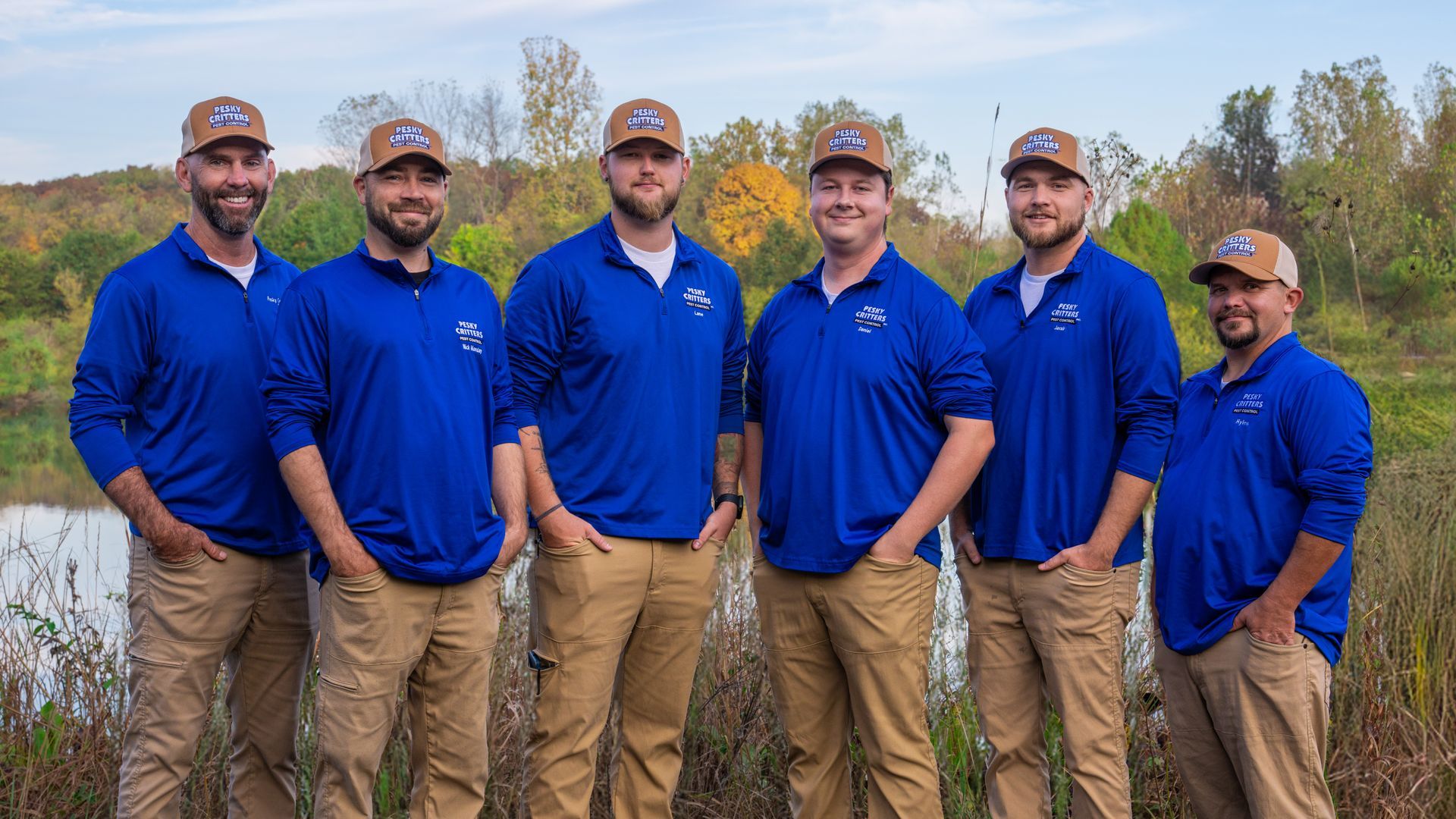 Six people in matching blue shirts and tan pants with hats stand together outdoors by a body of water.