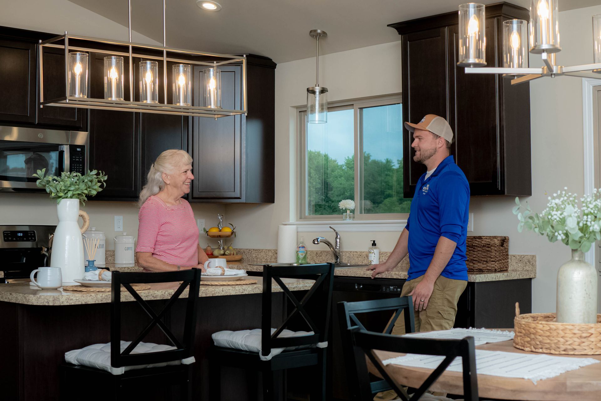 A worker in a blue shirt talking to a customer inside a modern kitchen with dark cabinets and a granite island.