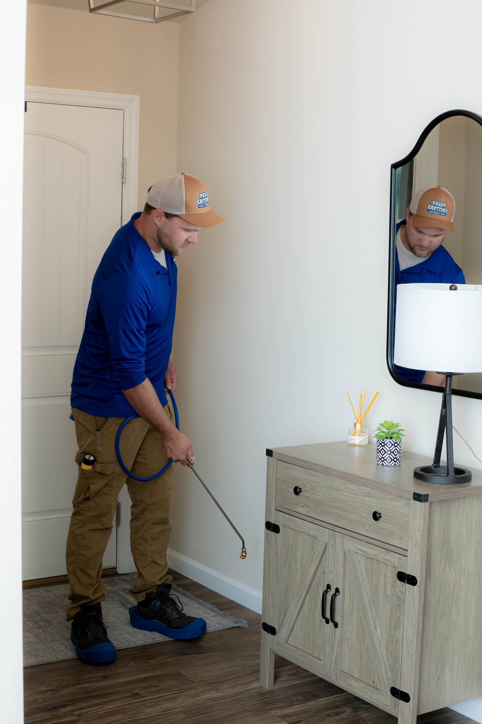 A technician in a blue shirt and cap uses a long-handled sprayer to treat the baseboards in a residential hallway.