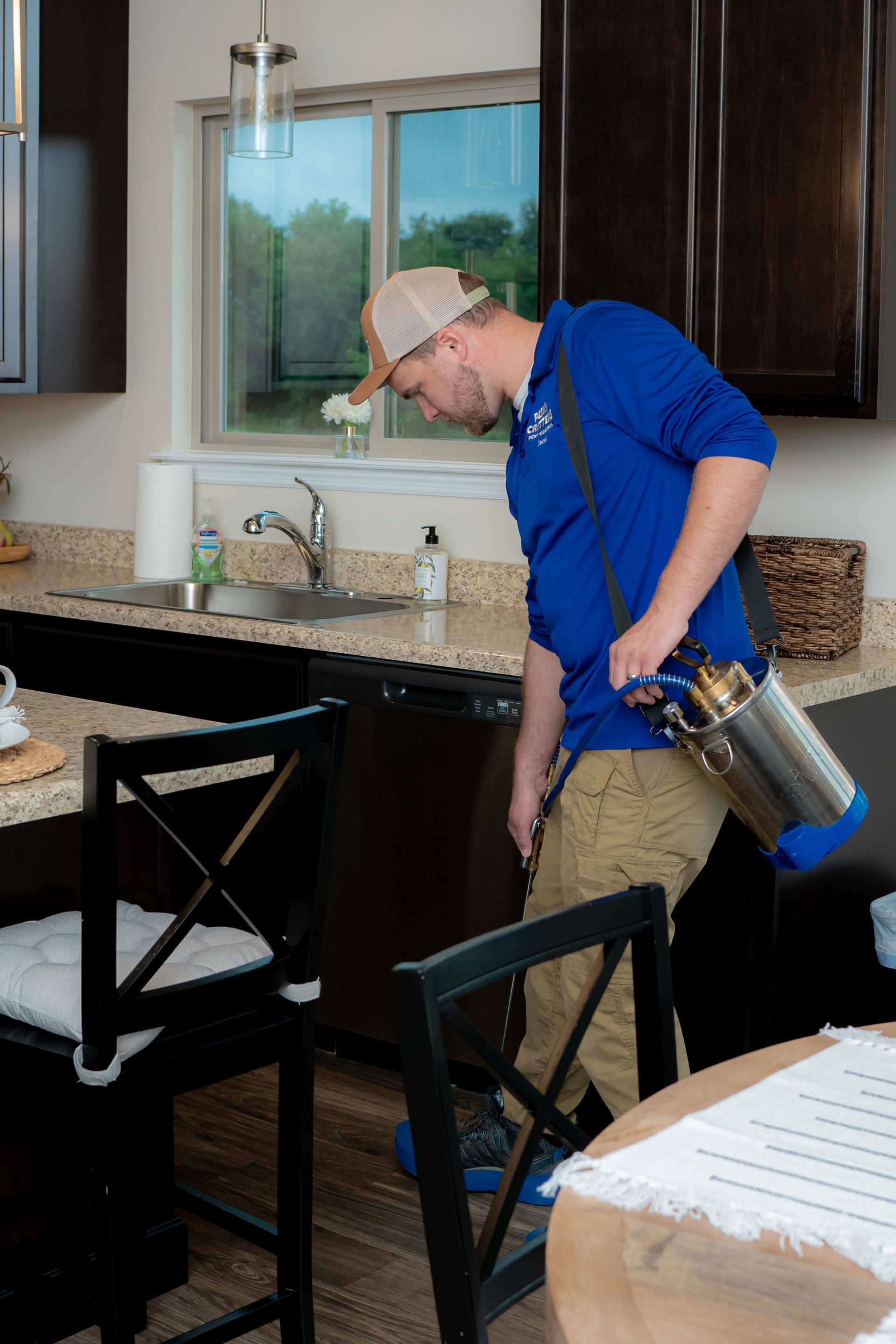 A pest control technician in a blue uniform sprays a kitchen floor near a dishwasher.
