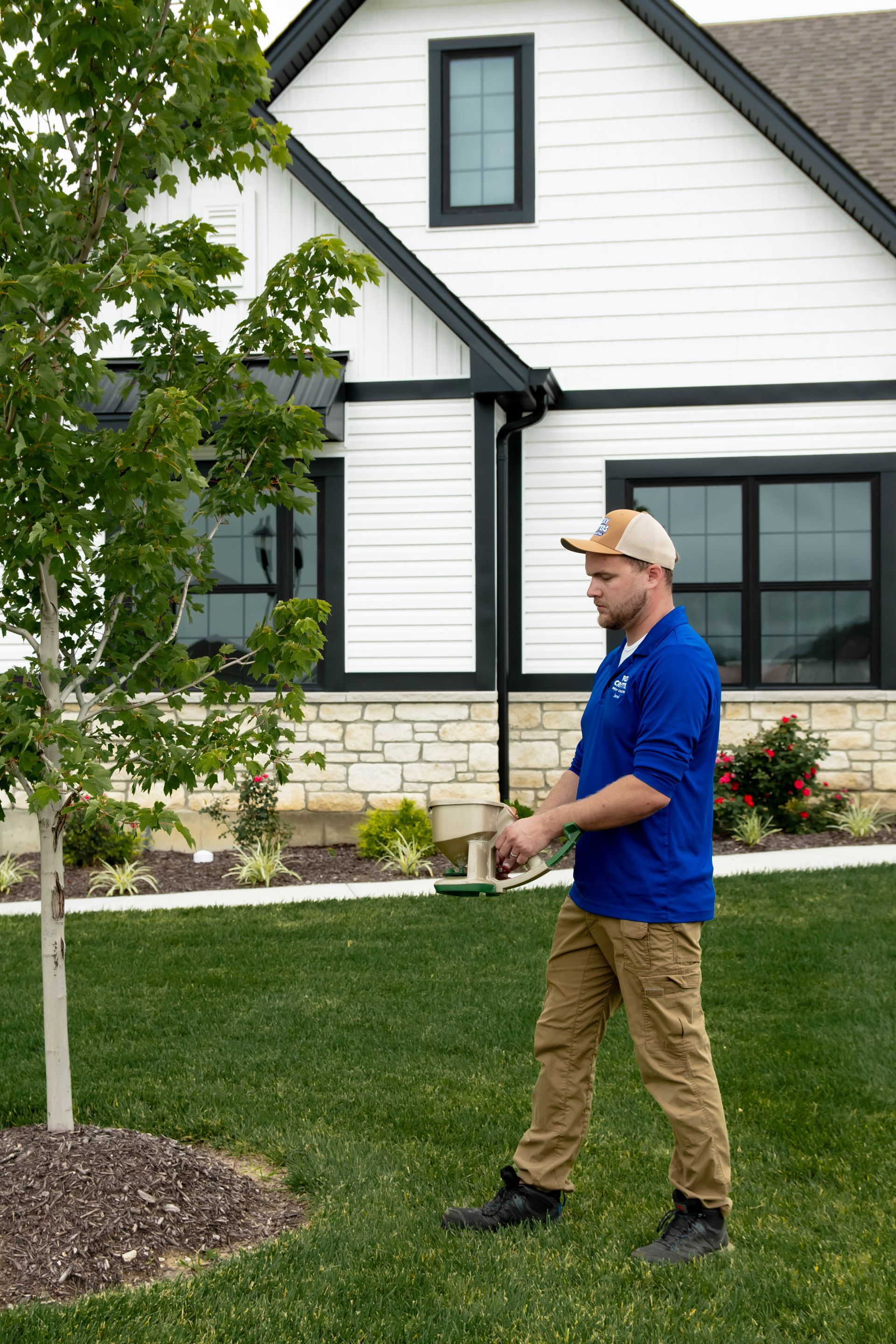 A person wearing a blue polo shirt and tan pants stands in a green yard near a young tree in front of a white house.