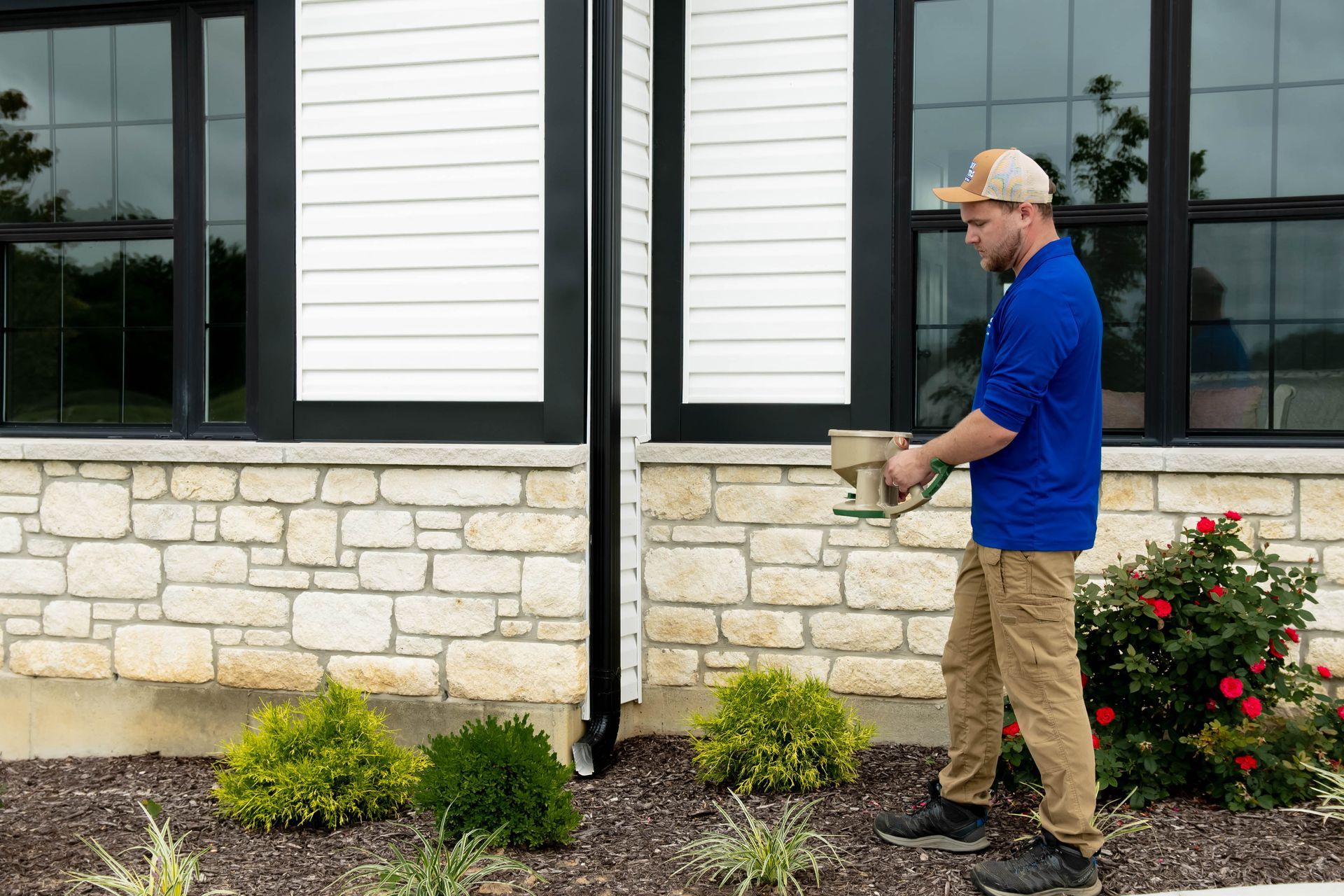 A person in a blue shirt and khaki pants walks alongside a building, applying pest control treatment to the foundation.