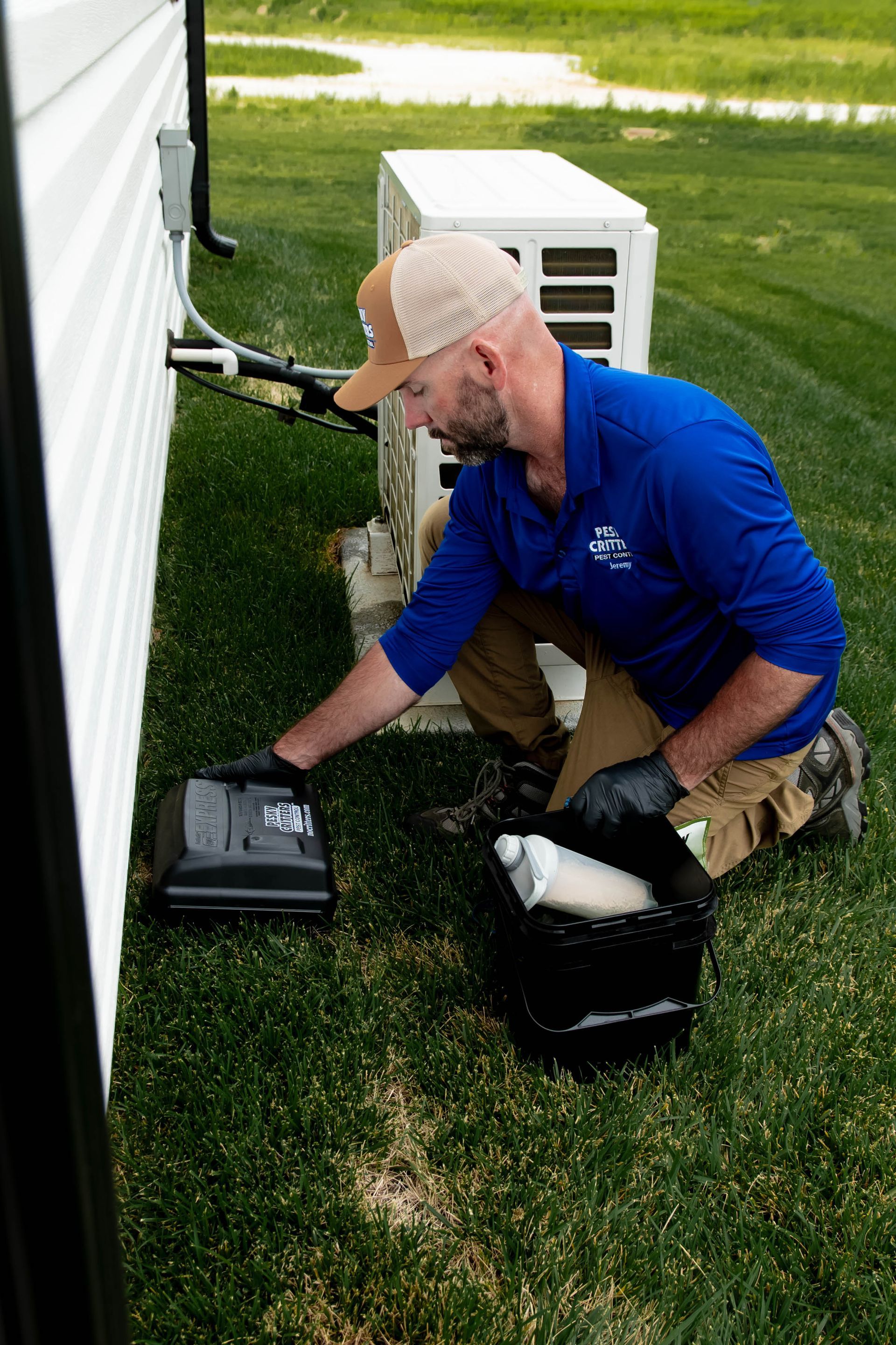 A technician in a blue shirt kneels outside, placing a pest control bait station near the exterior wall of a building.