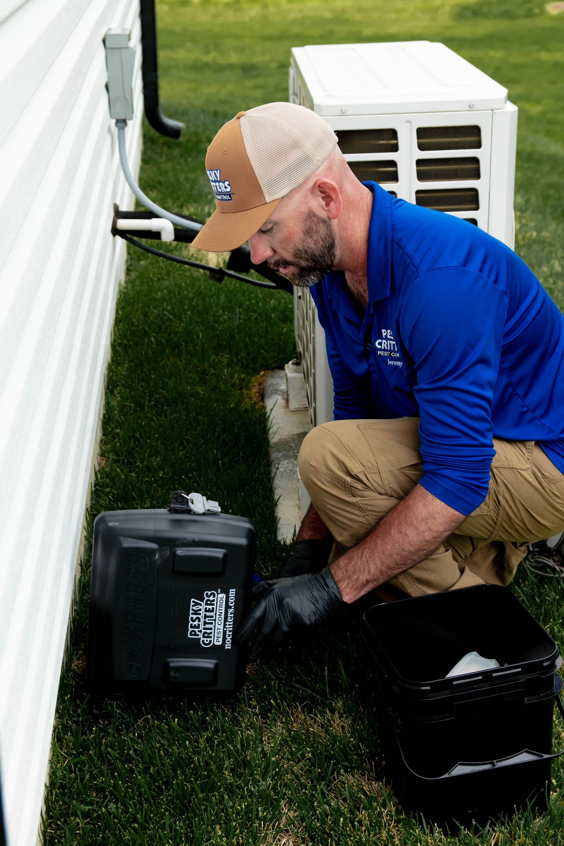 A technician wearing a blue shirt and cap crouches beside a home exterior to work on an HVAC unit with tools.