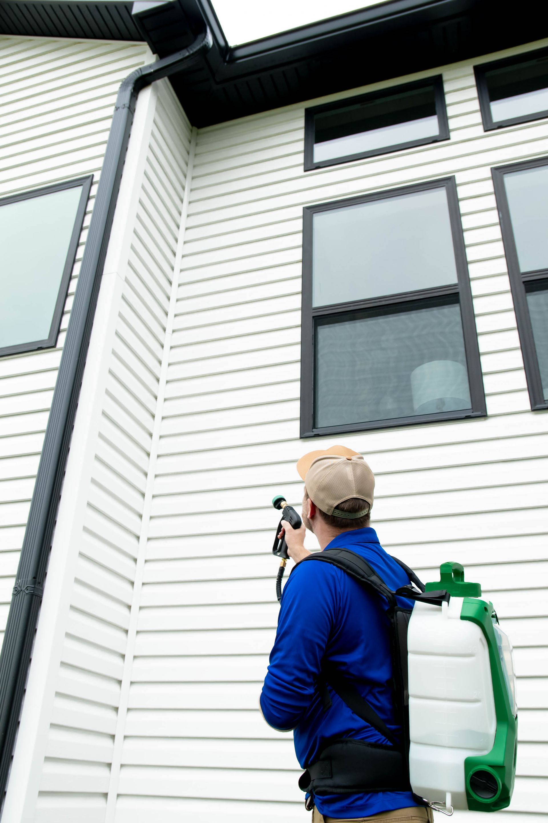A person wearing a blue shirt and backpack sprayer treats the exterior siding of a white house near large windows.