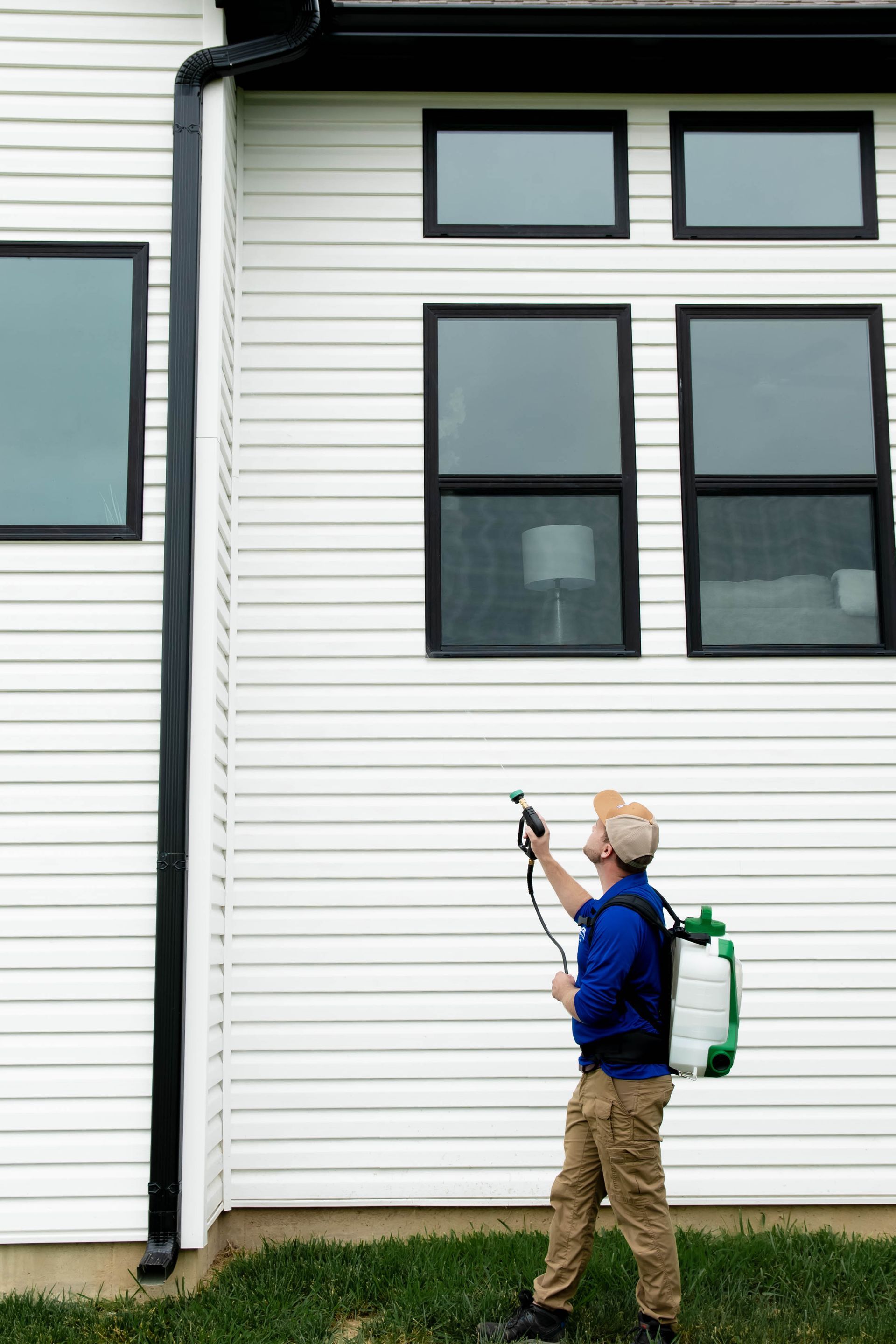 A person in a blue shirt and khaki pants uses a backpack sprayer to apply treatment to the exterior of a white house.