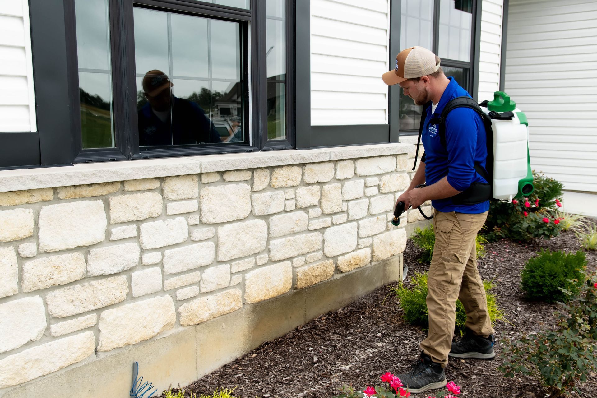 A pest control technician in a blue shirt sprays the foundation of a stone-faced house.