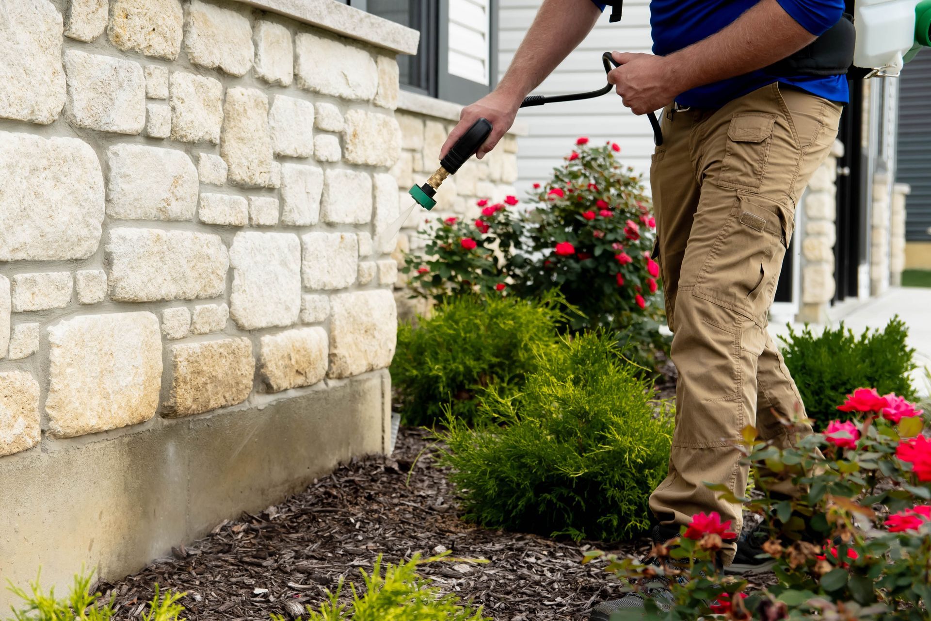 A pest control technician sprays a perimeter treatment against a stone-walled house foundation.