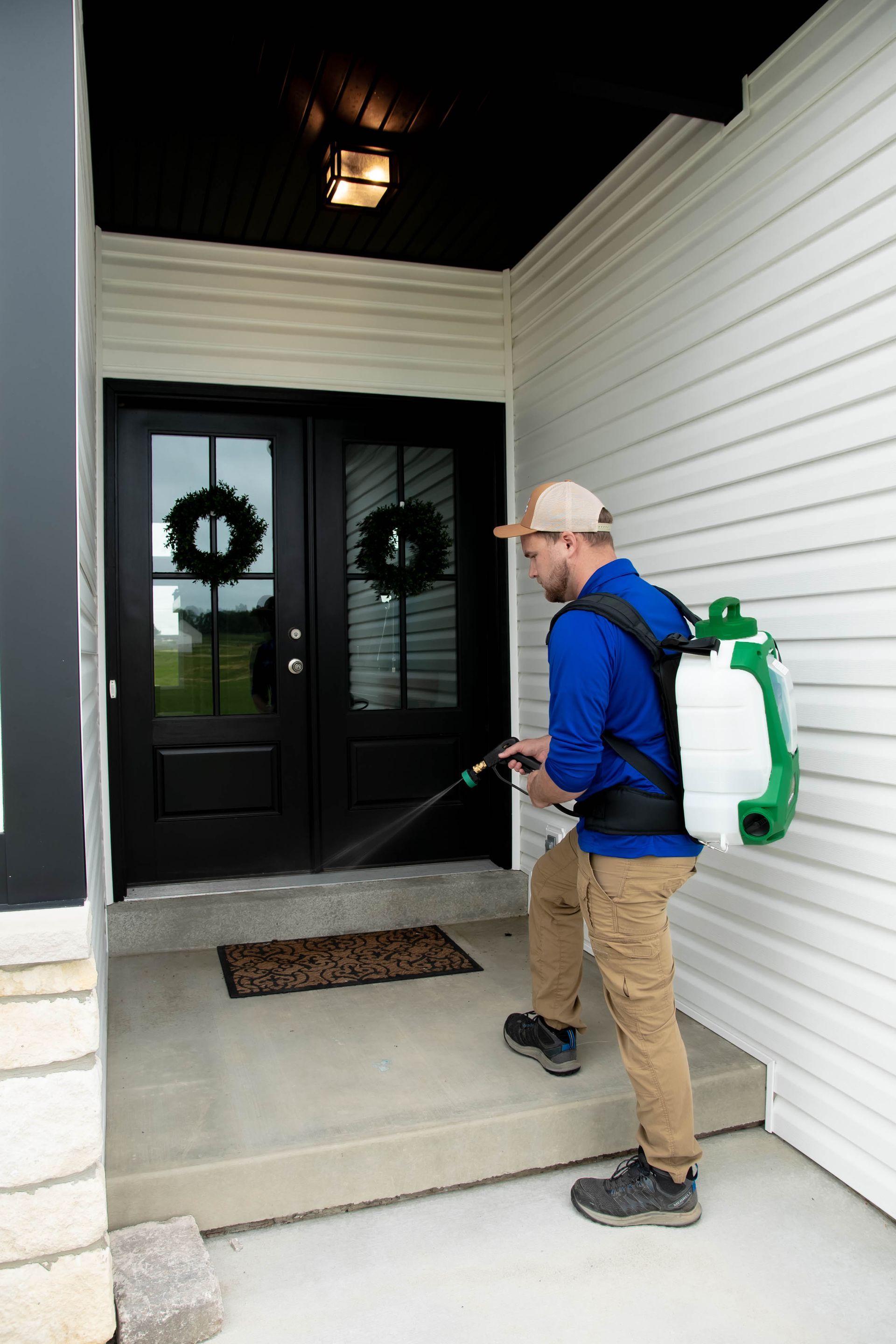 A person wearing a blue shirt and backpack sprayer applies treatment to the exterior of a home's front entryway.