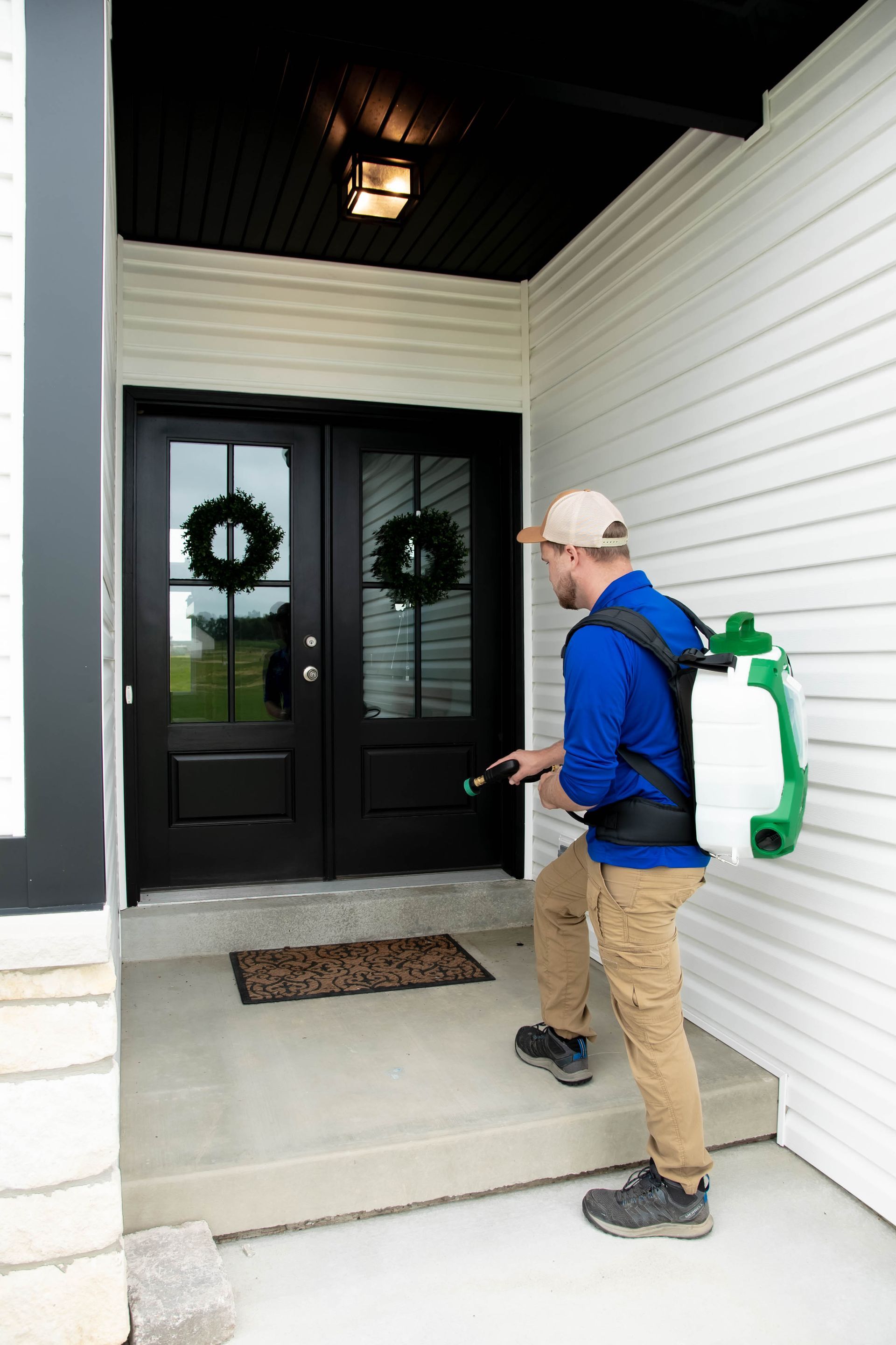 A person in a blue shirt and khaki pants wears a backpack sprayer while treating the doorway of a house.