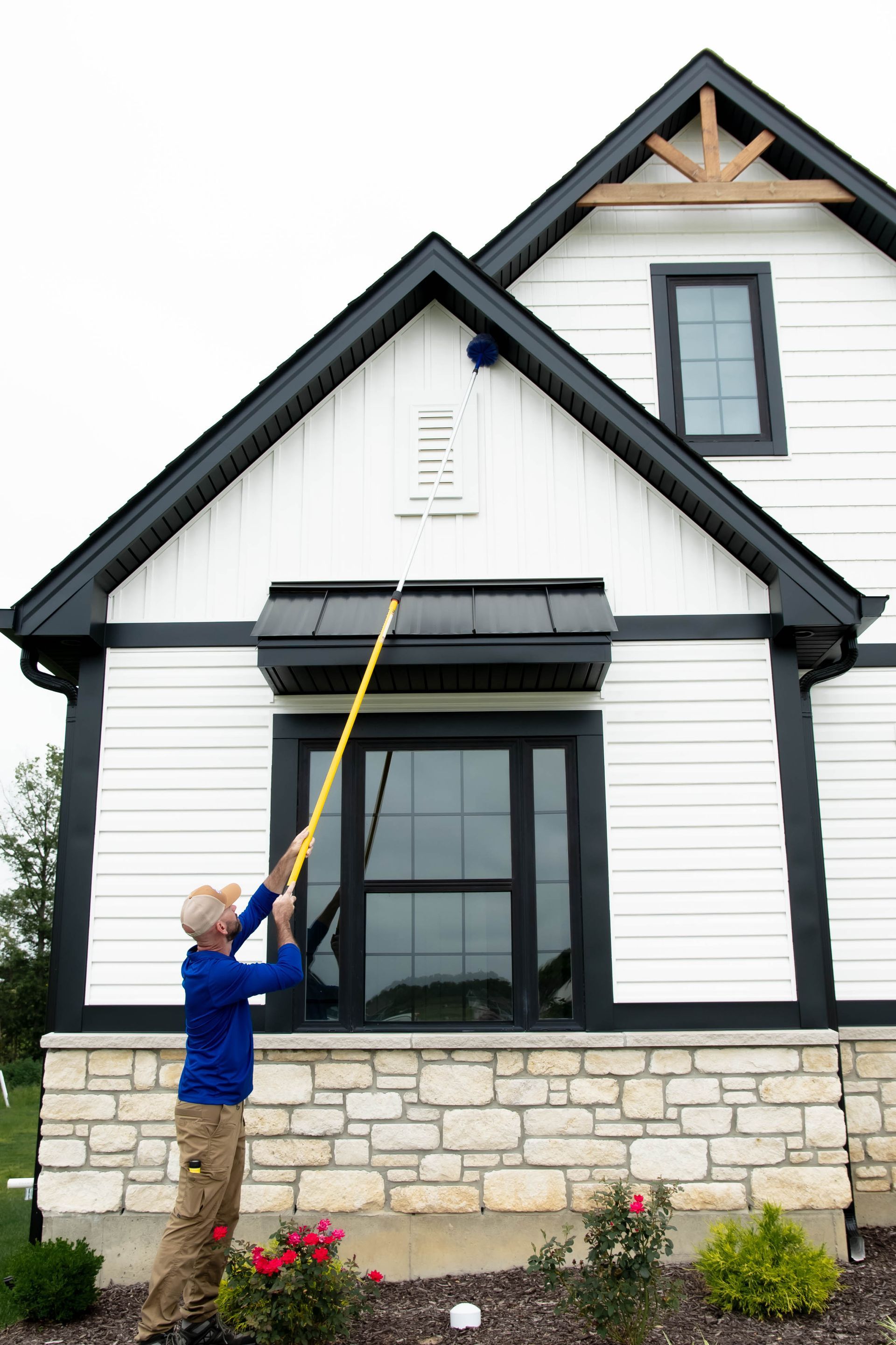 A person in a blue shirt uses an extended pole to clean the exterior siding of a white, modern home.