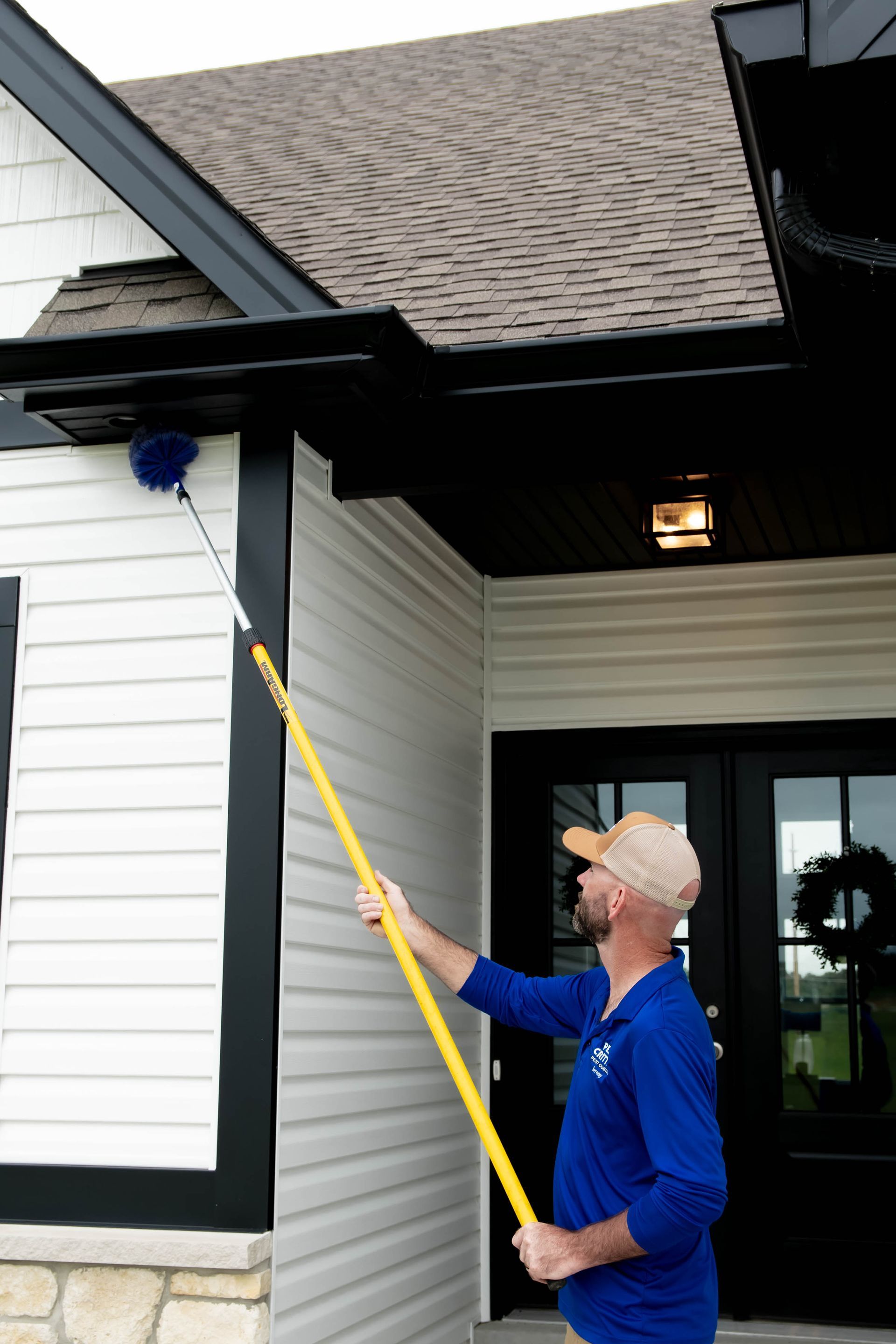 A person in a blue long-sleeved shirt uses a long yellow extension pole with a duster to clean the black eaves of a house.