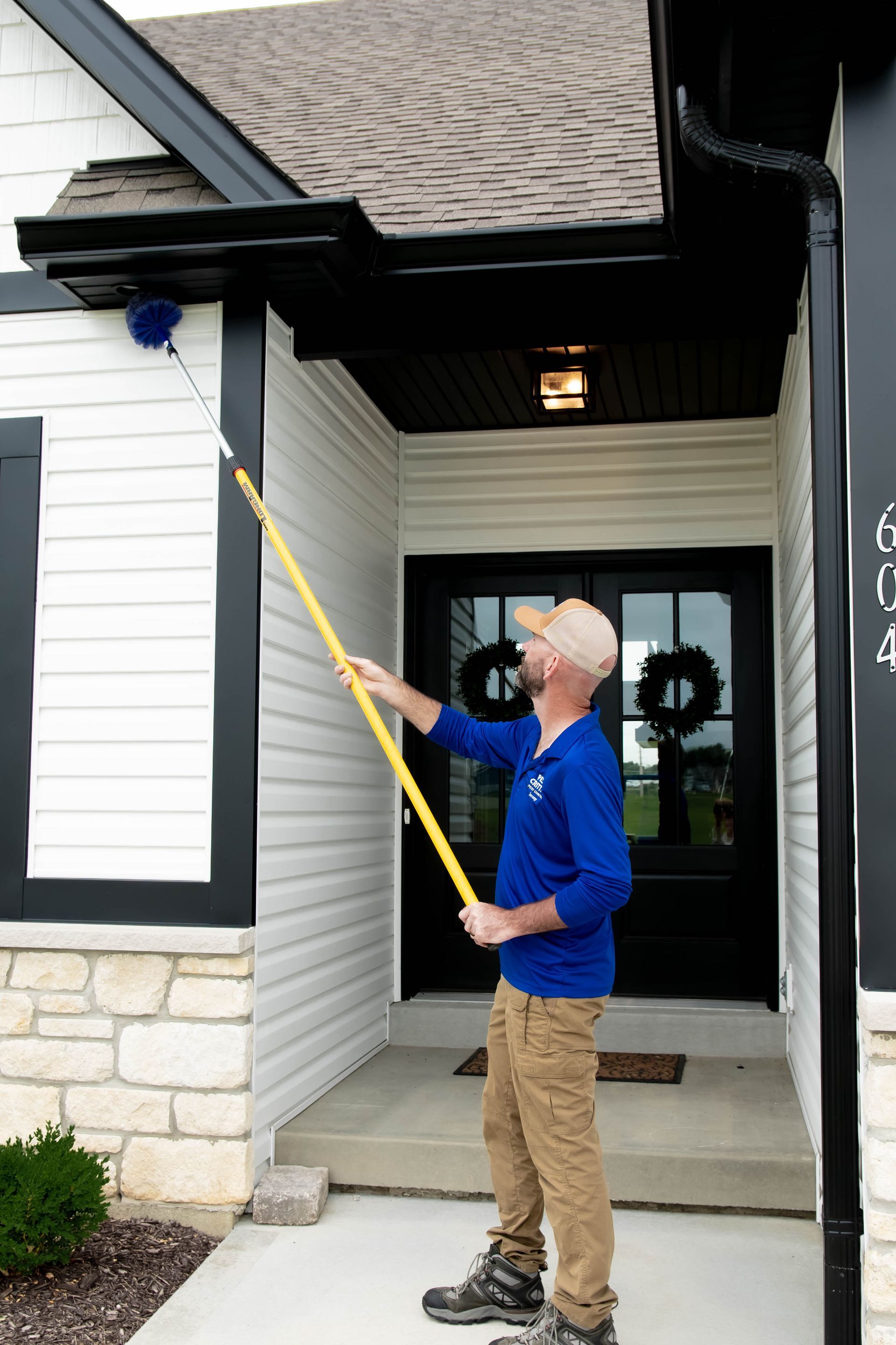 A person wearing a blue shirt uses an extended pole with a blue brush to clean the soffit of a white and black house.