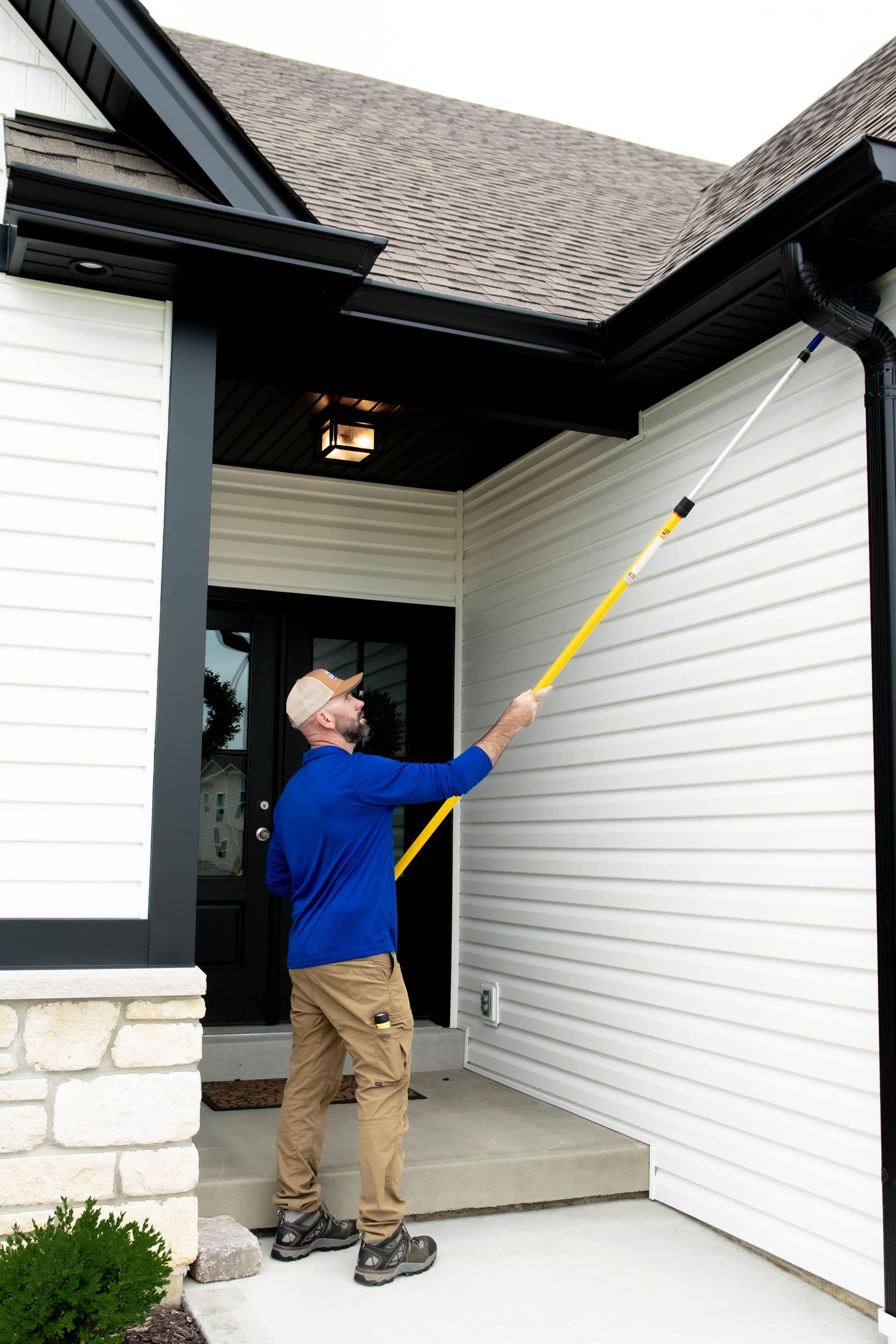 A person in a blue shirt uses a yellow extension pole to clean a black gutter on the exterior of a white house.