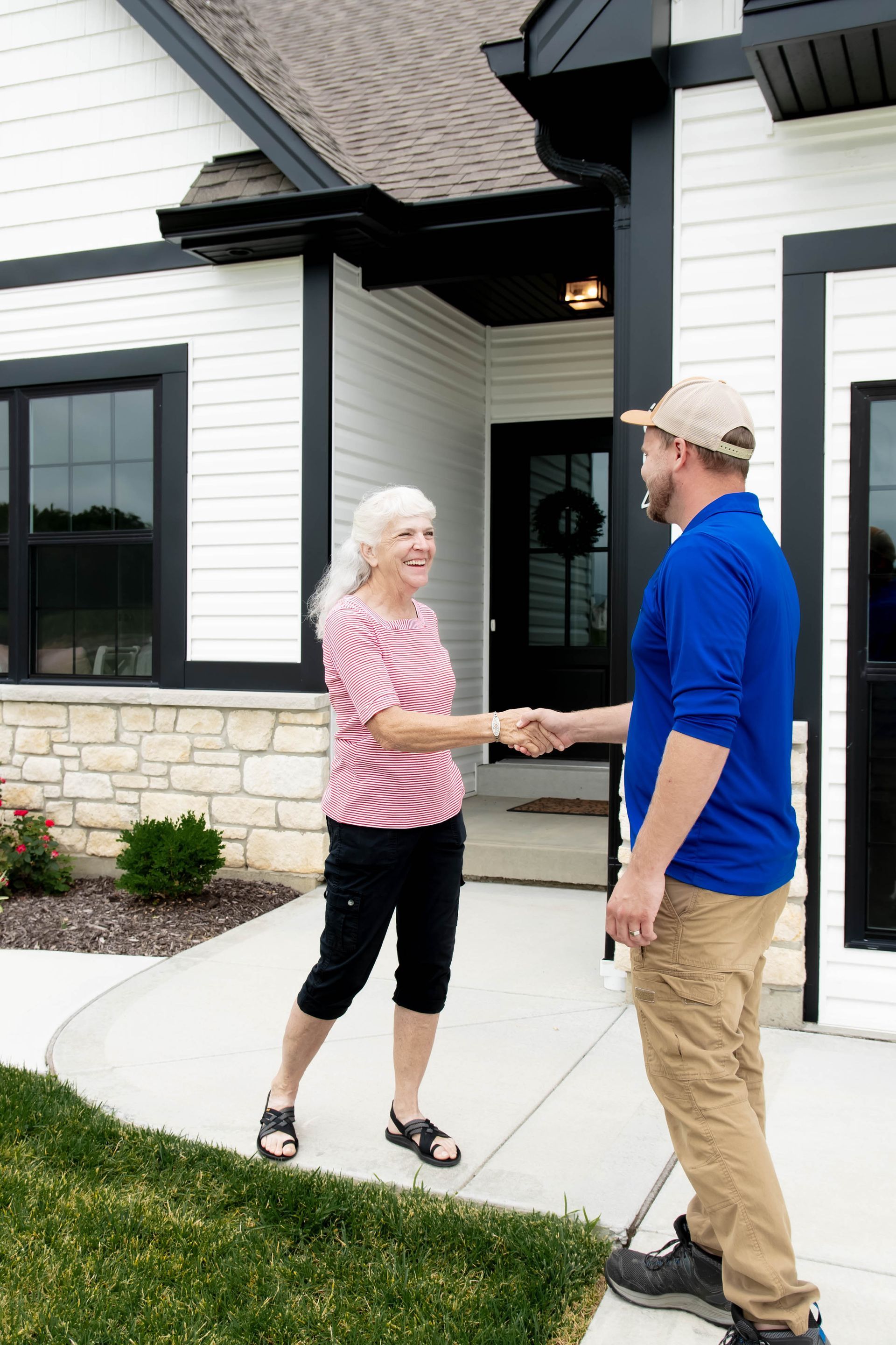 A smiling person in a red striped shirt shakes hands with a worker in a blue polo on the front porch of a house.