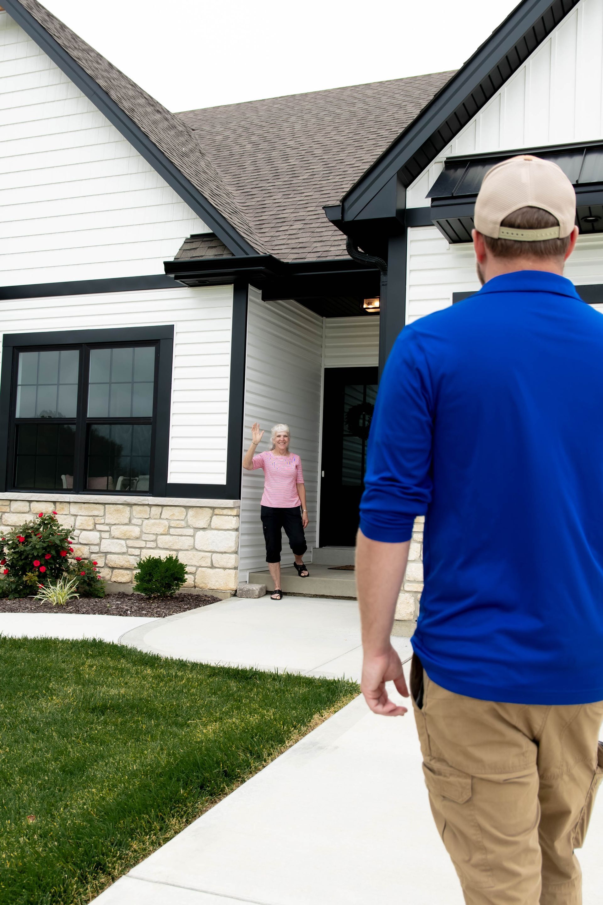 A person in a blue shirt walks toward a doorway where another person stands and waves in front of a white house.