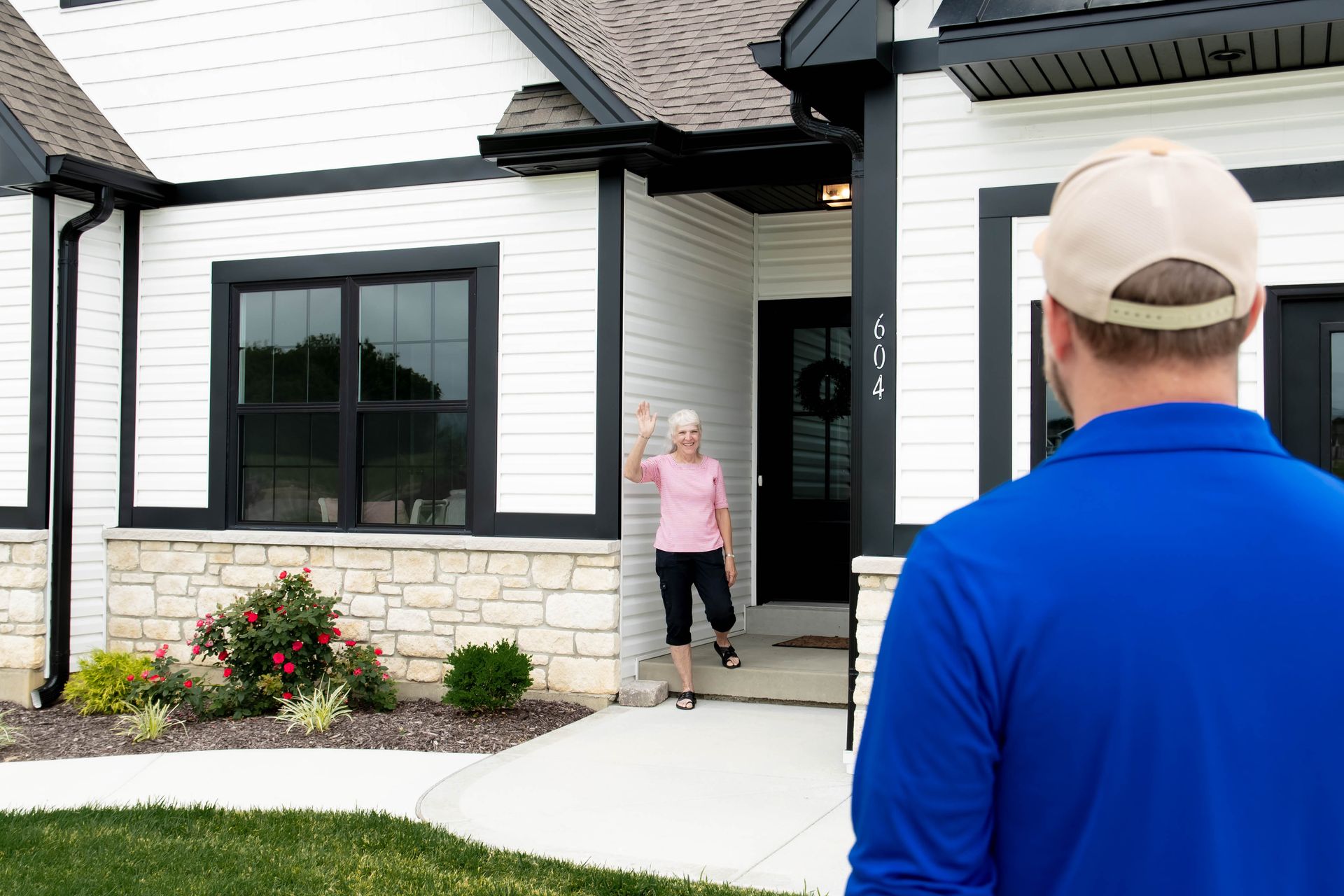 A person in a blue shirt and hat stands before a house while a person in the doorway waves at them.