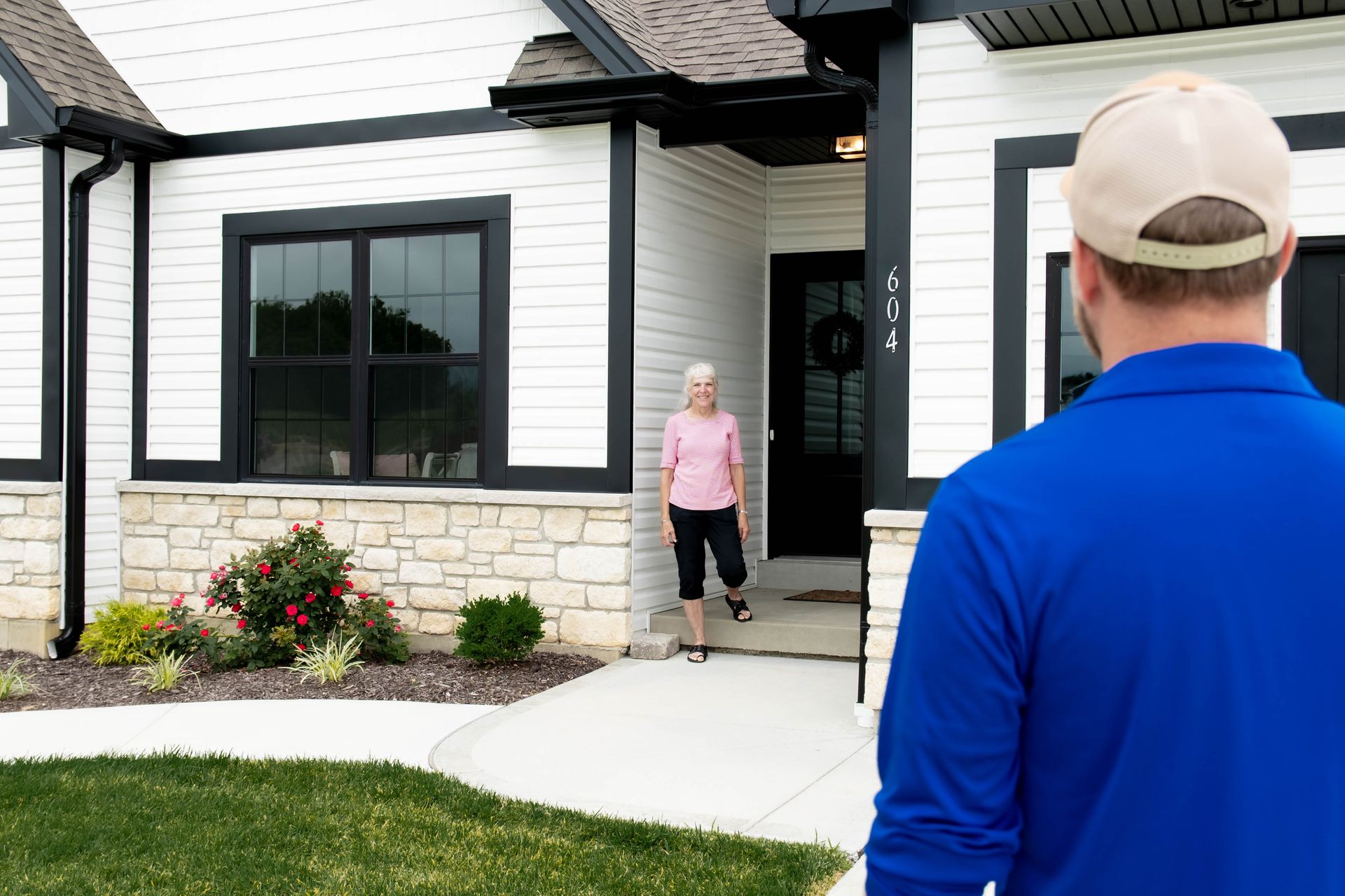 A person in a blue shirt and baseball cap approaches the front door of a white house, where a person stands in the doorway.