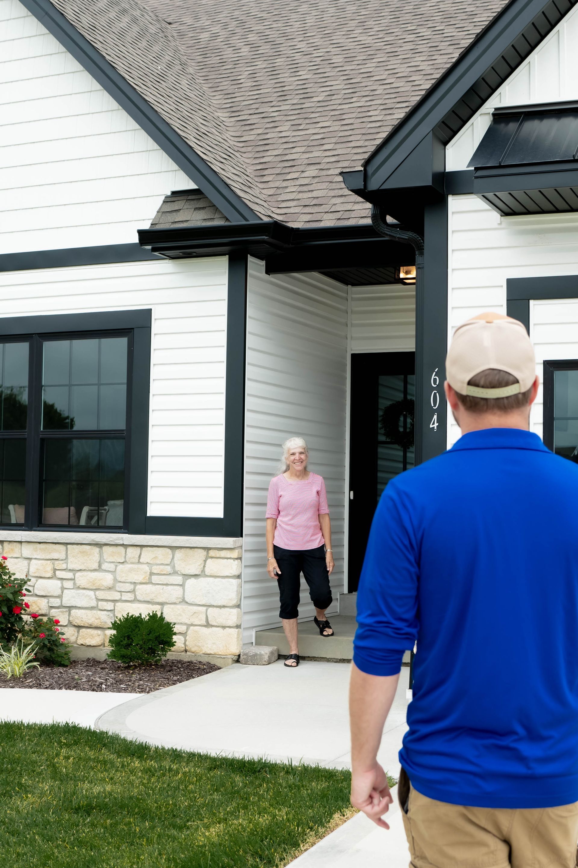 A person in a blue shirt walks toward a doorway where an individual stands in a modern white house with stone trim.