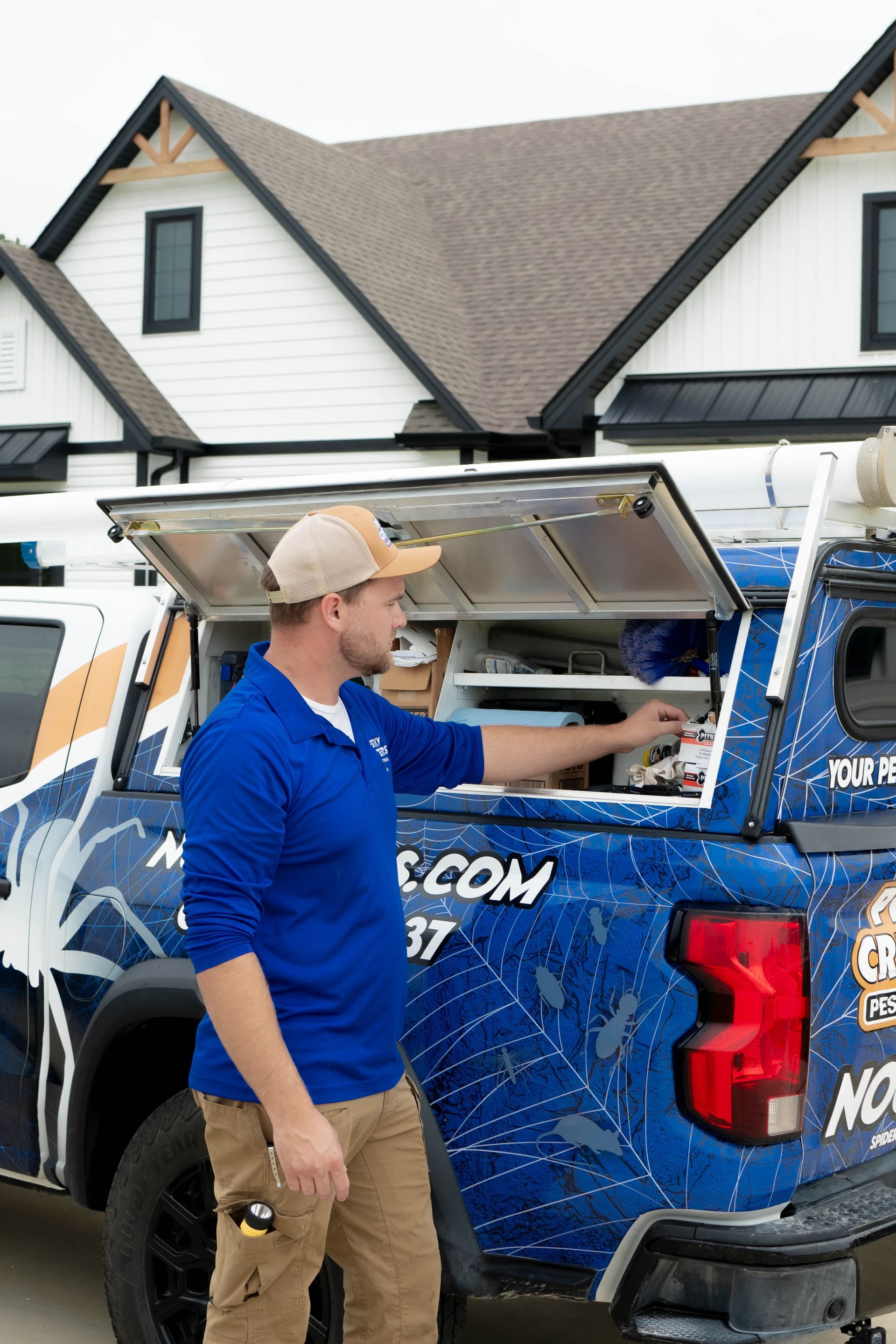 A technician in a blue polo and tan cap retrieves equipment from the open storage compartment of a pest control truck.