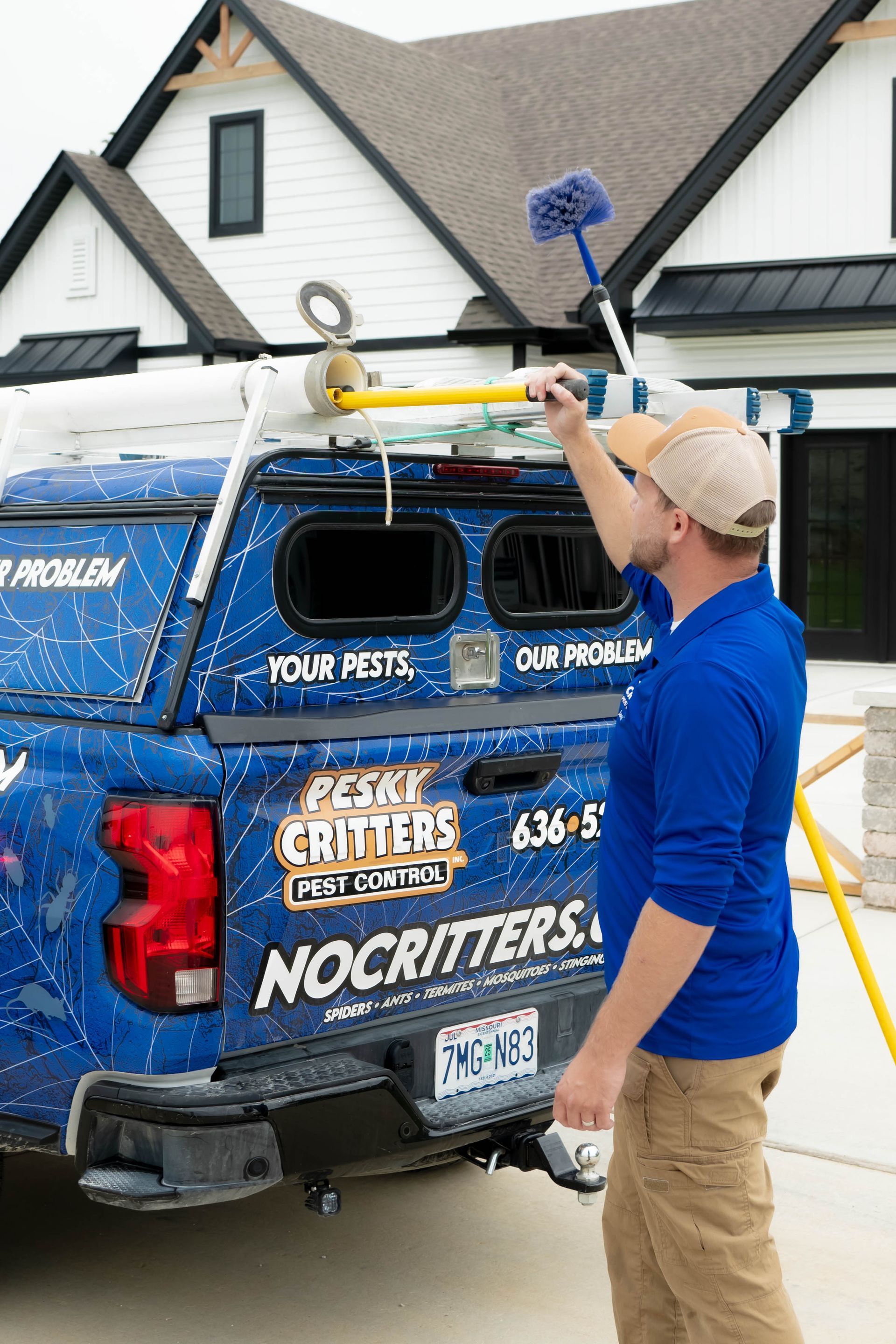 A person in a blue shirt and tan pants retrieves a long pole from a blue pest control service truck parked by a house.