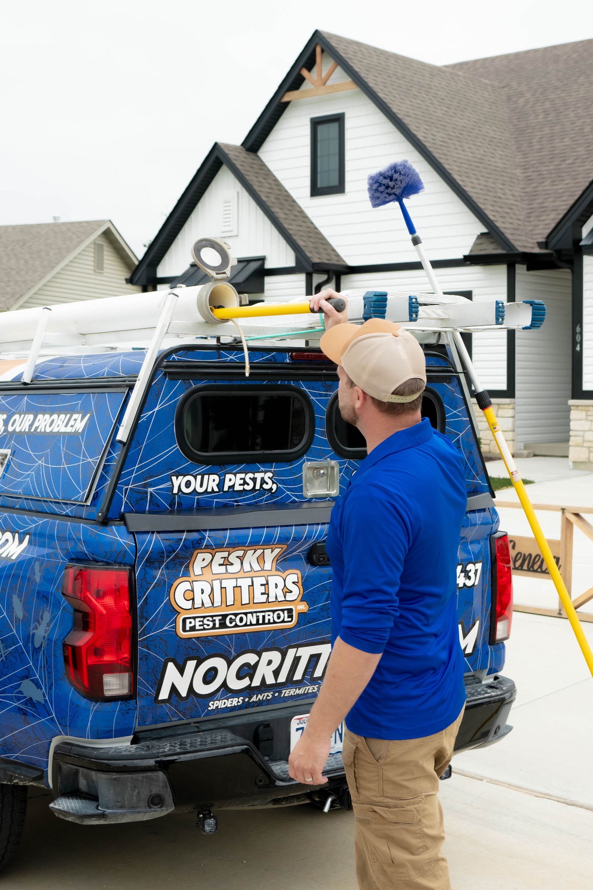 A pest control technician in a blue shirt works with equipment stored on the rack of a blue branded truck near a house.