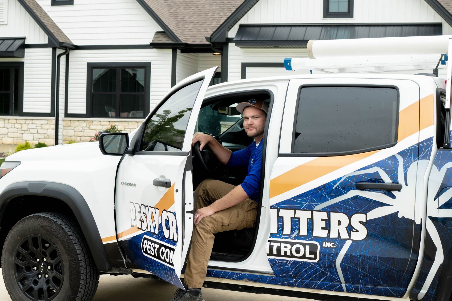 A service technician sits in the open driver's door of a white pest control truck parked in front of a house.