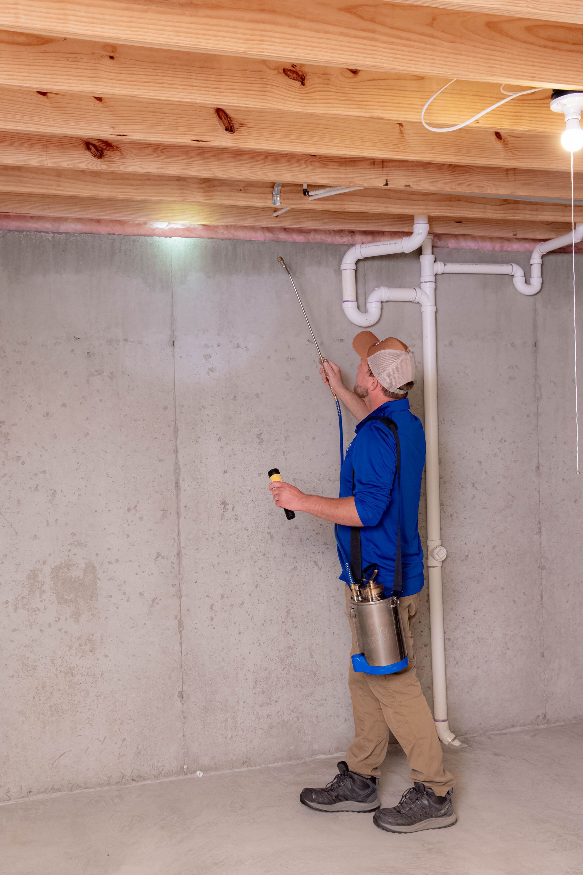 A person in a blue shirt and khaki pants uses a spray wand to treat a concrete basement wall.