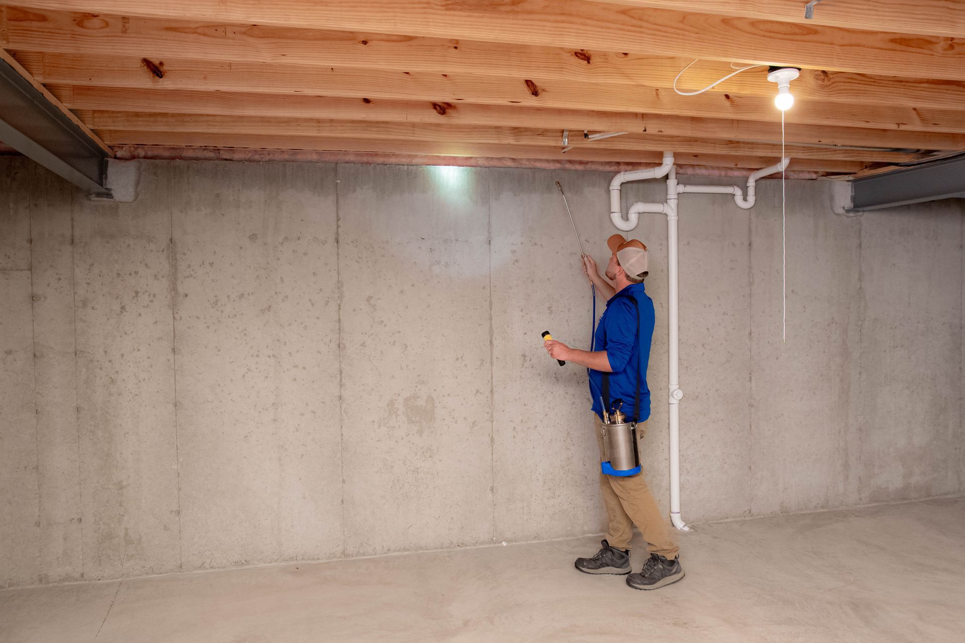A person in a blue shirt sprays a sealant on a bare concrete basement wall.